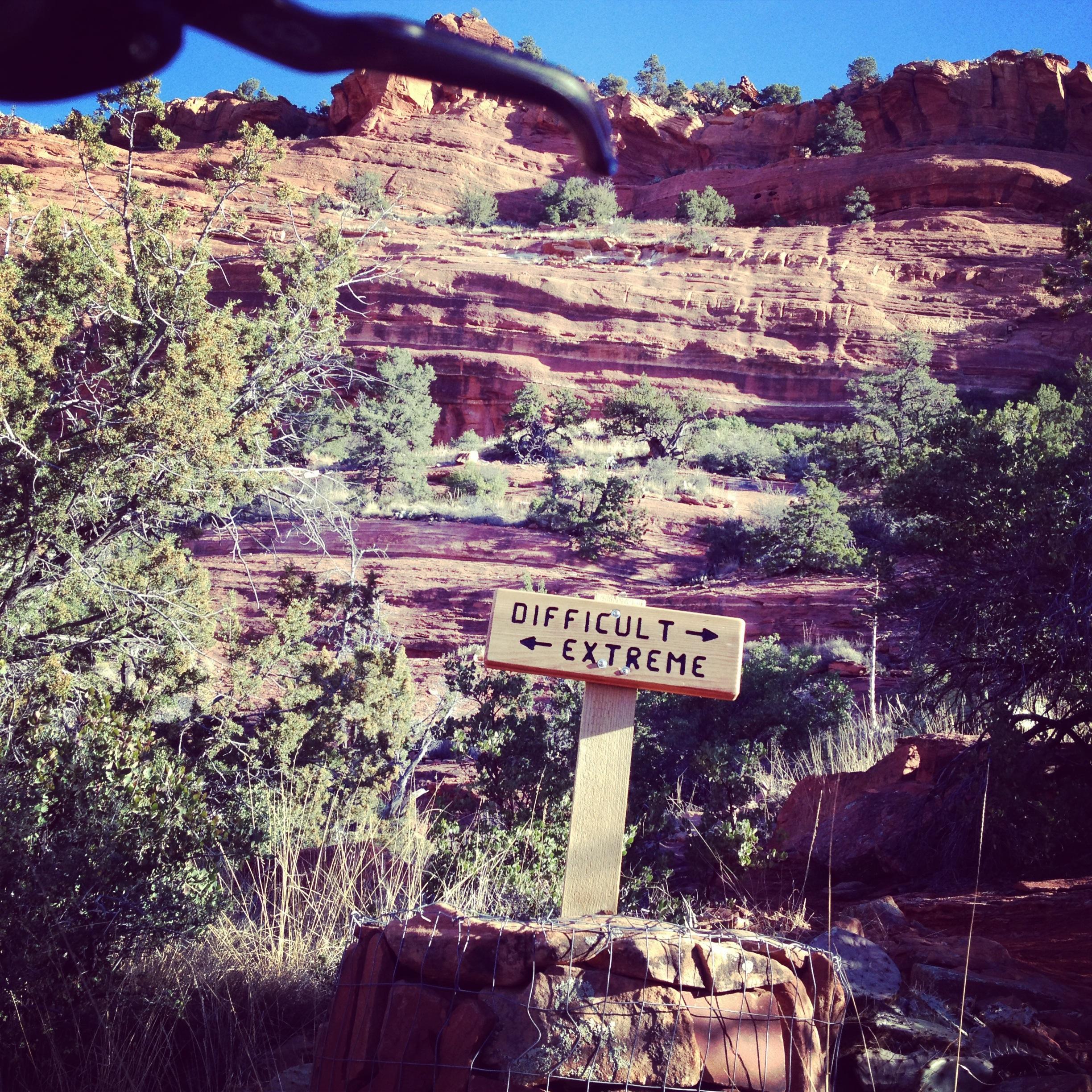 A wooden signpost in a natural landscape displaying directions for hiking difficulty levels, with the words "Difficult" and "Extreme" pointed in opposite directions. Behind the sign is a backdrop of red rock formations and greenery under a blue sky. Mescal Trail mountain bike trail.