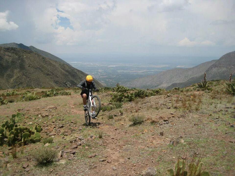 A mountain biker performs a wheelie on a dirt path surrounded by rocky terrain and cacti, with a panoramic view of valleys and distant mountains under a cloudy sky. Real de Catorce Trails mountain bike trail.