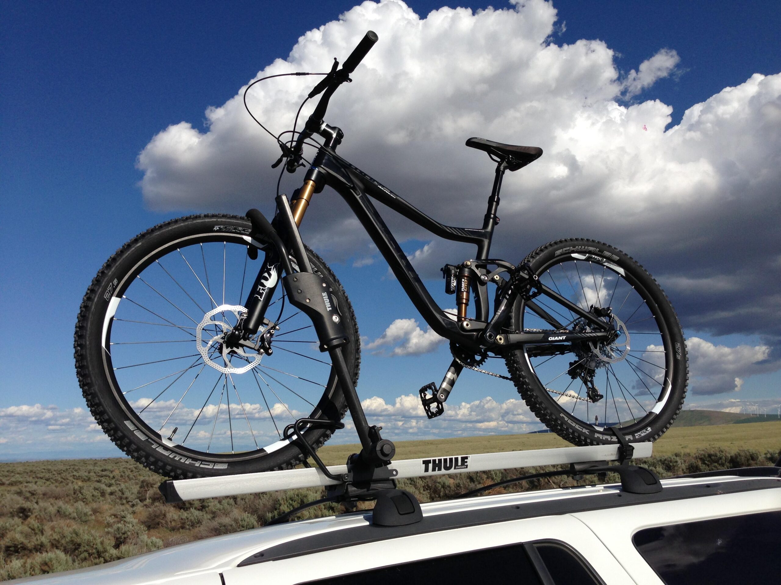 Giant Trance Advanced SX 27.5: A black mountain bike is securely mounted on the roof rack of a vehicle. The background features a clear blue sky with fluffy white clouds, and a landscape of sagebrush and rolling hills is visible below.