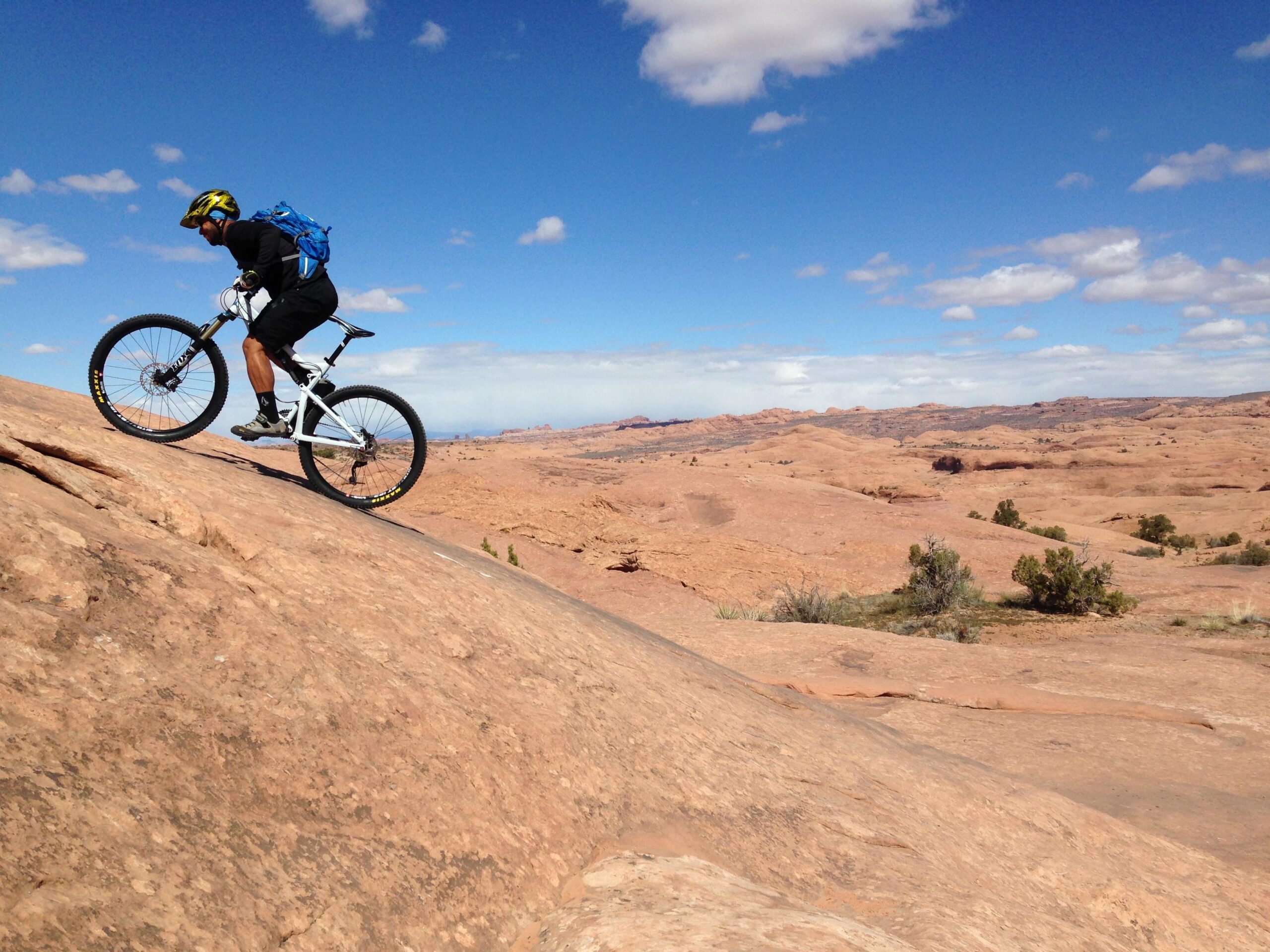 A mountain biker navigating a rocky terrain under a clear blue sky, with expansive desert landscape in the background and scattered vegetation. The cyclist is wearing a helmet and a backpack, demonstrating an adventurous outdoor activity. Slickrock mountain bike trail.