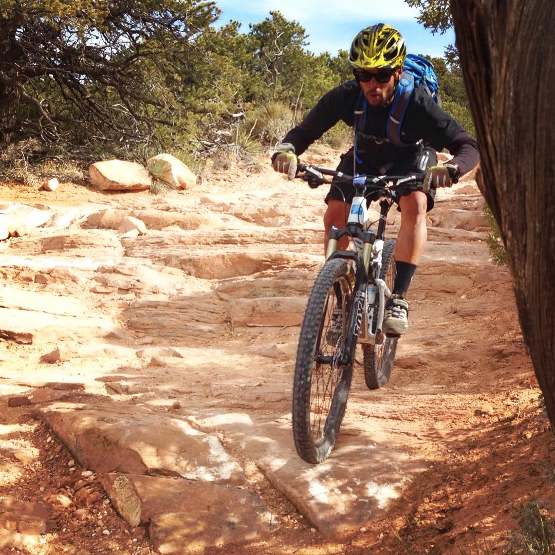 A mountain biker navigates a rocky trail, pedaling uphill on a sunny day. The cyclist is wearing a helmet, sunglasses, and a backpack, showcasing an active outdoor adventure in a natural landscape. Porcupine Rim mountain bike trail.