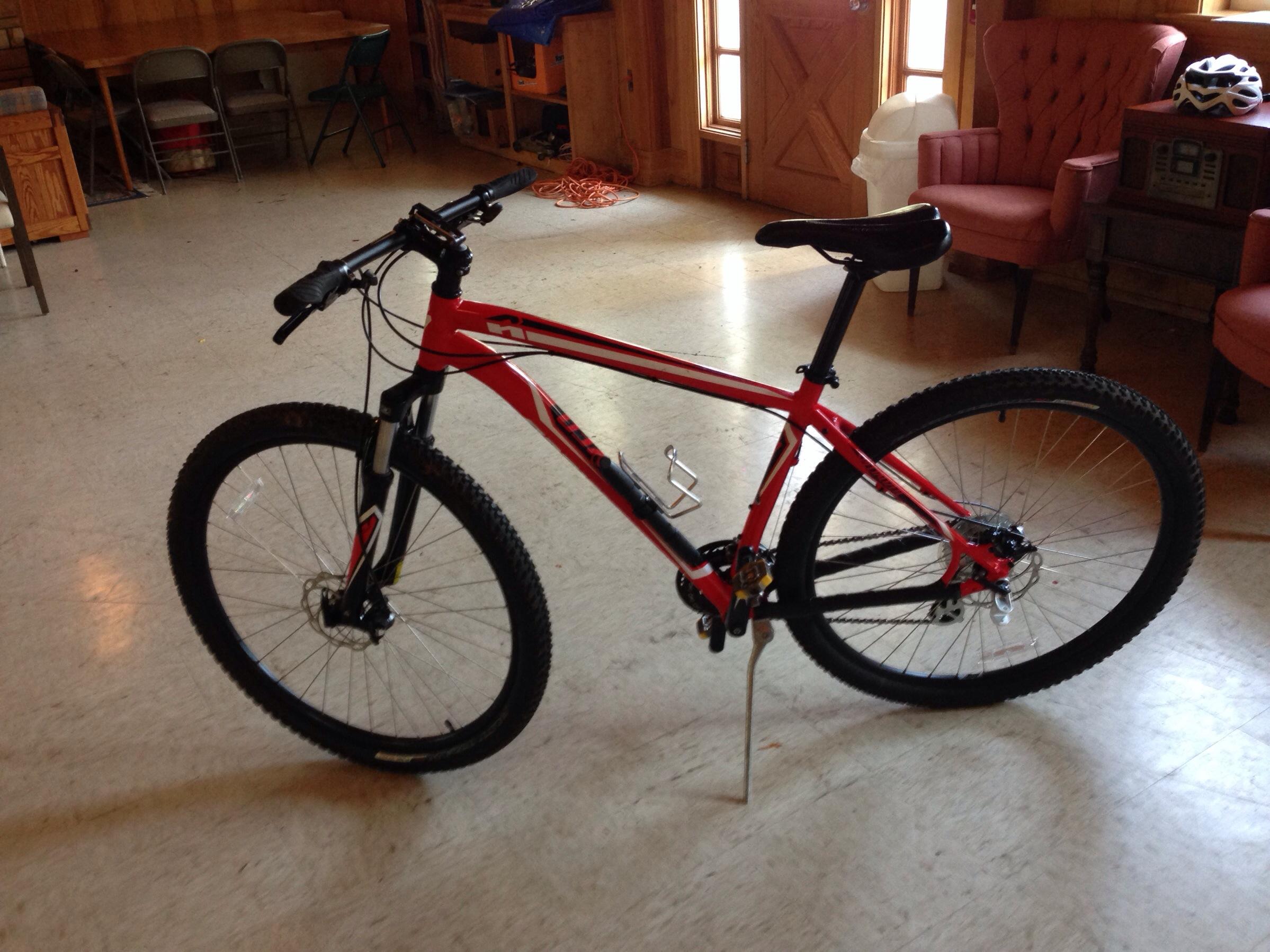 Specialized Hardrock Disc 29: A red mountain bike with black accents is positioned in an indoor space featuring wooden walls and various pieces of furniture. The bike is standing on a light-colored floor, with a pair of rose-colored chairs visible in the background along with a wooden table and a retro radio.
