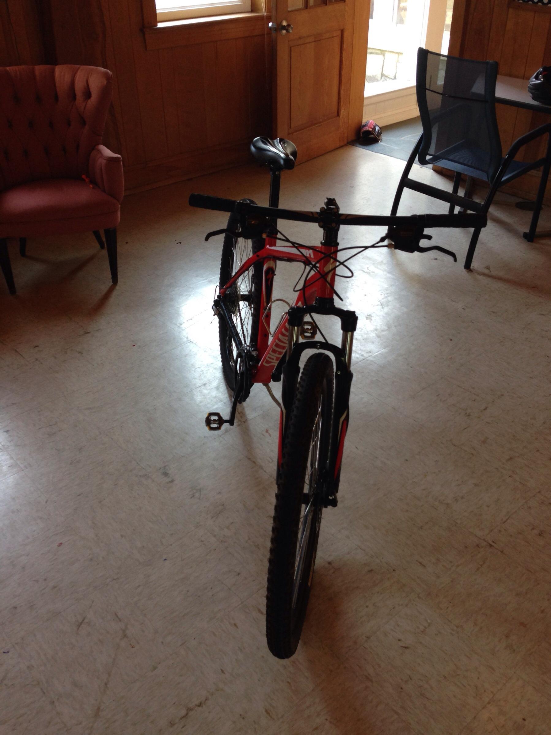 Specialized Hardrock Disc 29: A red mountain bike stands upright indoors on a light-colored floor. In the background, there is a pink upholstered chair and a black folding chair near a table. Natural light filters in through a window, illuminating the scene.