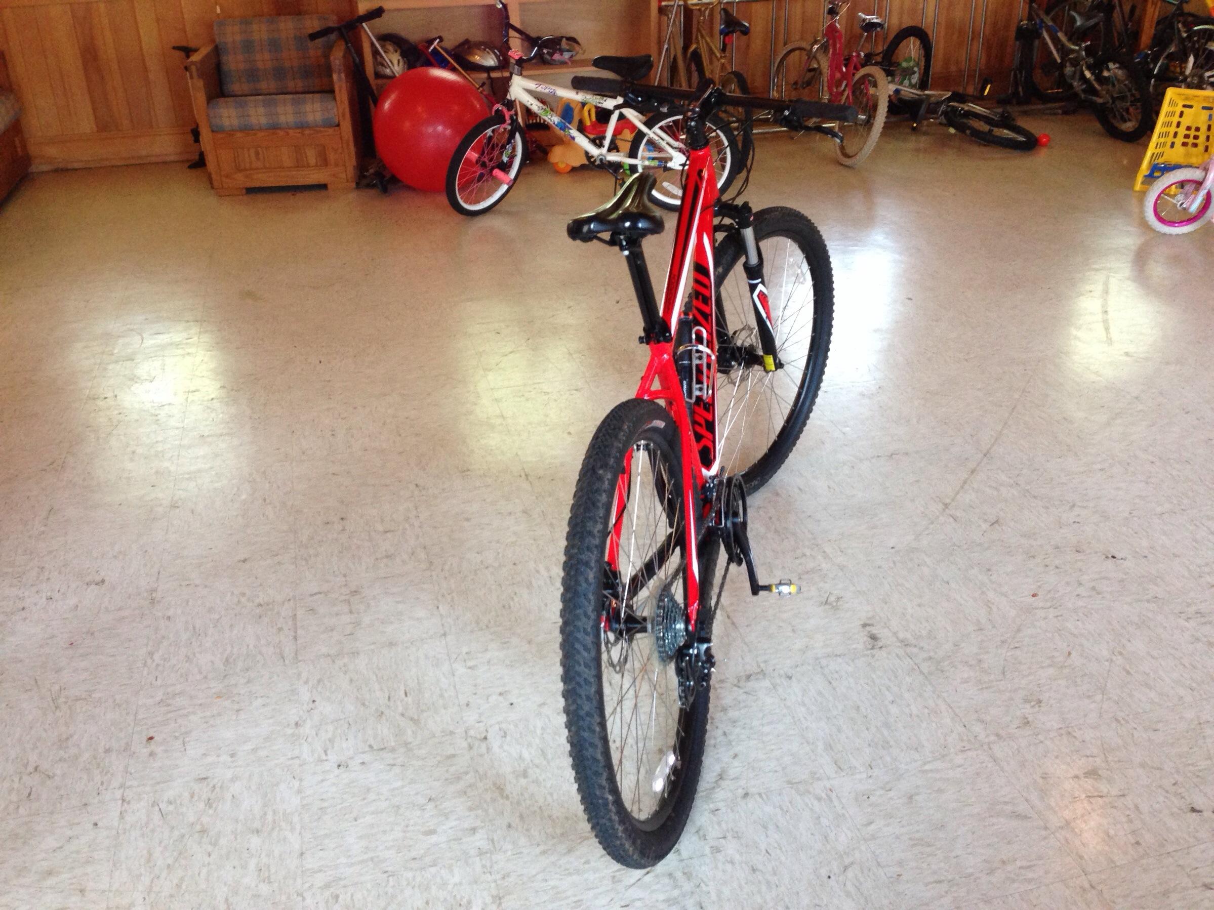 Specialized Hardrock Disc 29: A red mountain bike stands on a light-colored floor inside a room filled with various bicycles and play items, including a large red ball. Wooden walls and a couch are visible in the background.