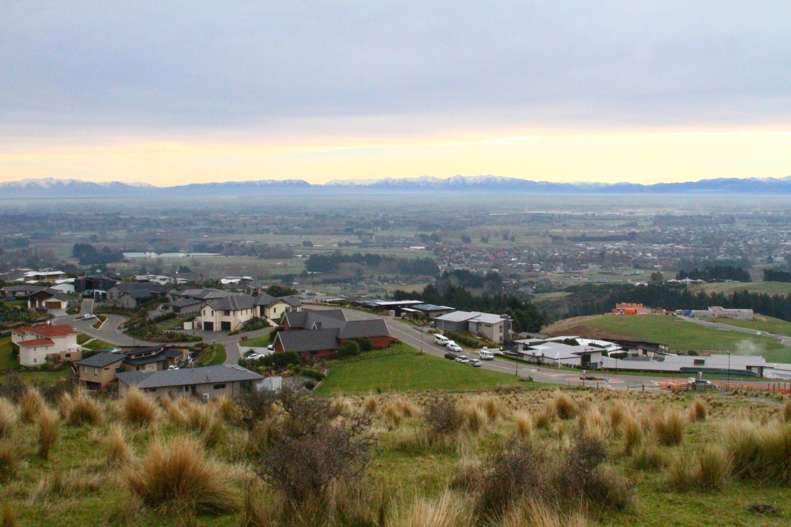 A panoramic view of a suburban area with various houses set on grassy hills. In the foreground, there are several residential buildings and winding roads, while the background features a vast landscape with fields and distant snow-capped mountains under a cloudy sky. Port Hills Tracks mountain bike trail.