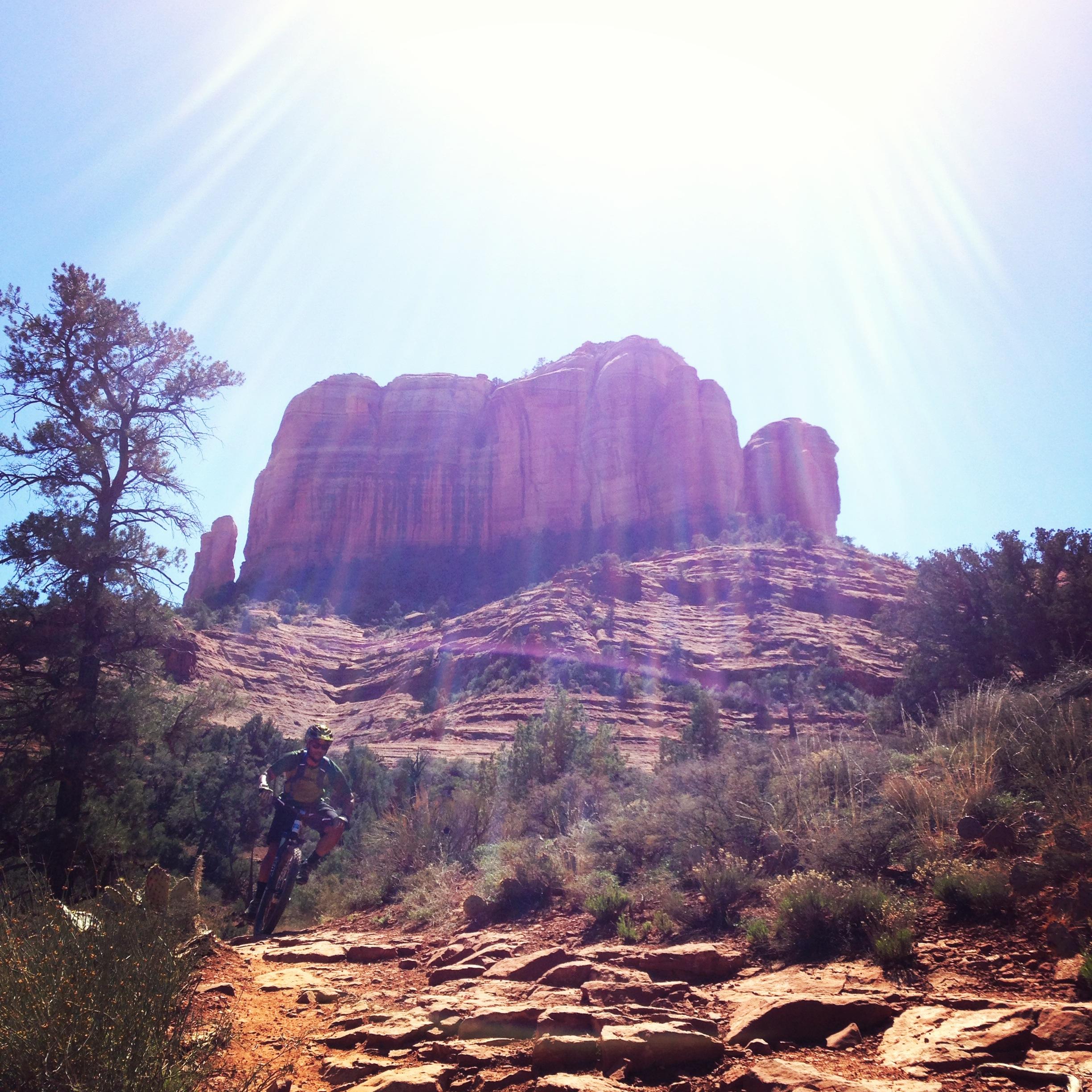 A cyclist riding on a rocky trail in a desert landscape, with a large red rock formation towering in the background under a bright sun. Trees and shrubs are visible along the trail. Templeton mountain bike trail.