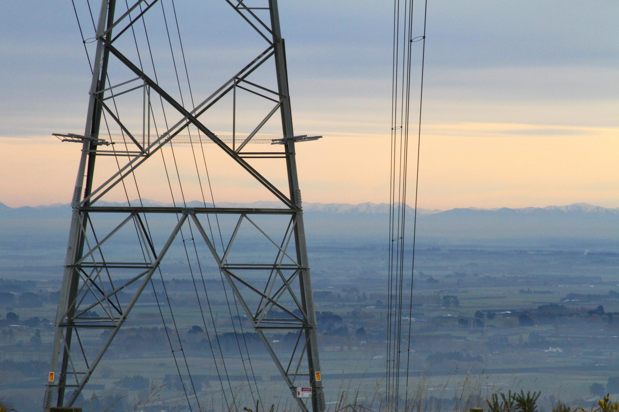 A tall metal transmission tower stands in the foreground, with power lines extending from it. In the background, a vast landscape of rolling hills and fields is visible, fading into a misty horizon with snow-capped mountains. The sky is softly illuminated in pastel hues as the sun sets. Port Hills Tracks mountain bike trail.