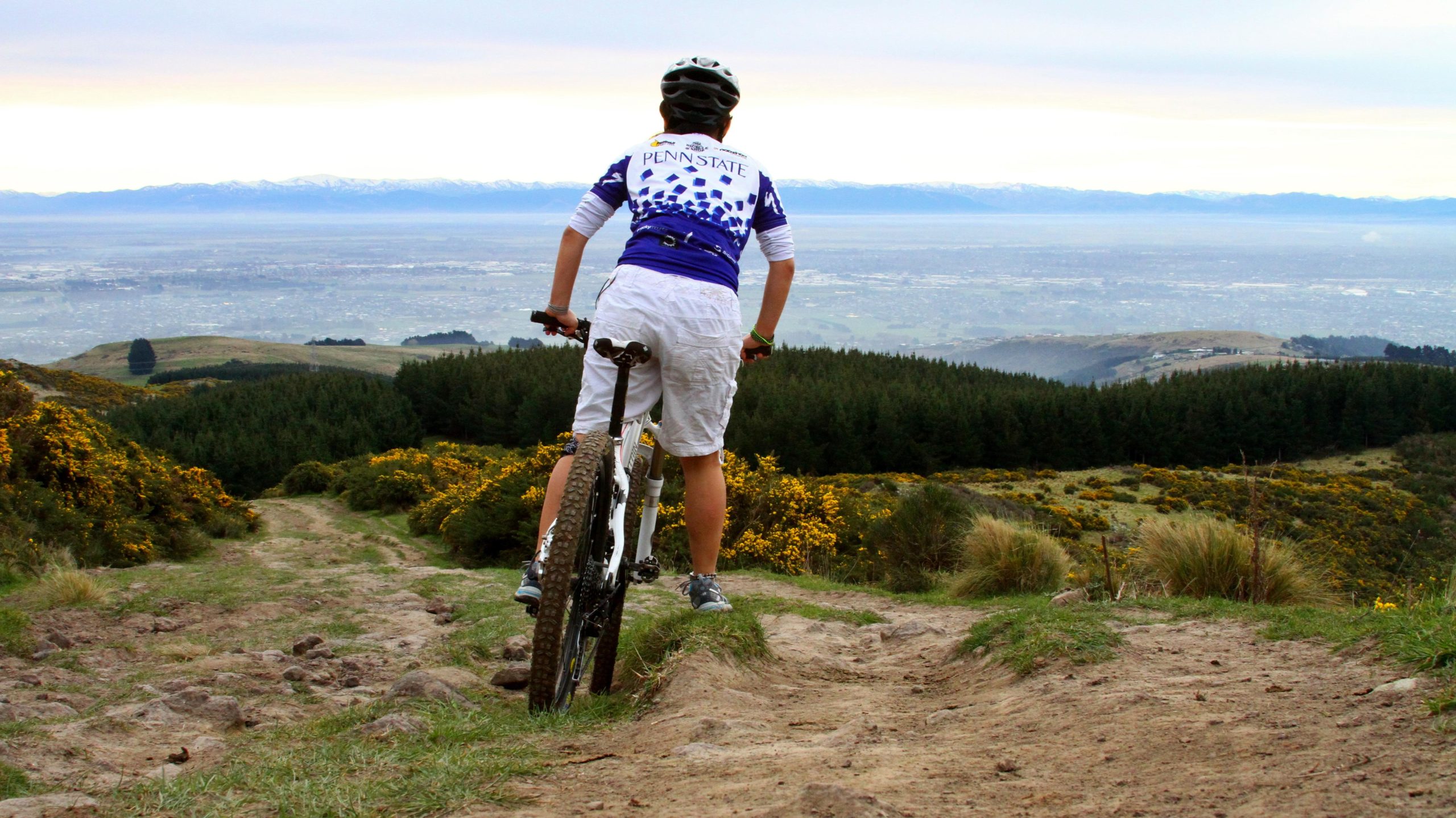 A person wearing a cycling jersey and shorts stands on a mountain biking trail, overlooking a panoramic view of a valley with distant mountains. The path is rocky and surrounded by greenery, with shrubs and yellow flowers in the foreground. Port Hills Tracks mountain bike trail.