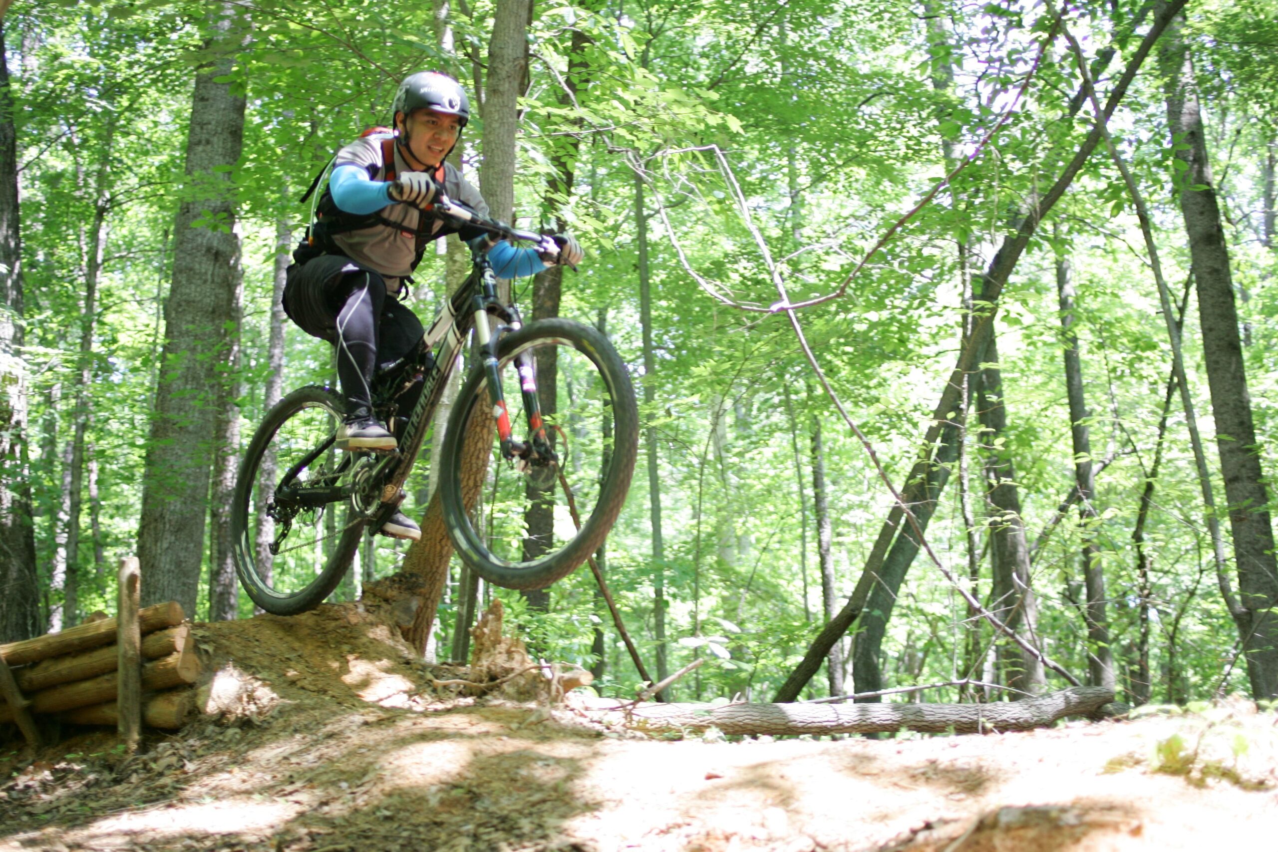 A mountain biker in a helmet performs a jump over a wooden ramp on a dirt trail surrounded by lush green trees. Sunlight filters through the foliage, creating a vibrant and energetic atmosphere. Salem Lake mountain bike trail.