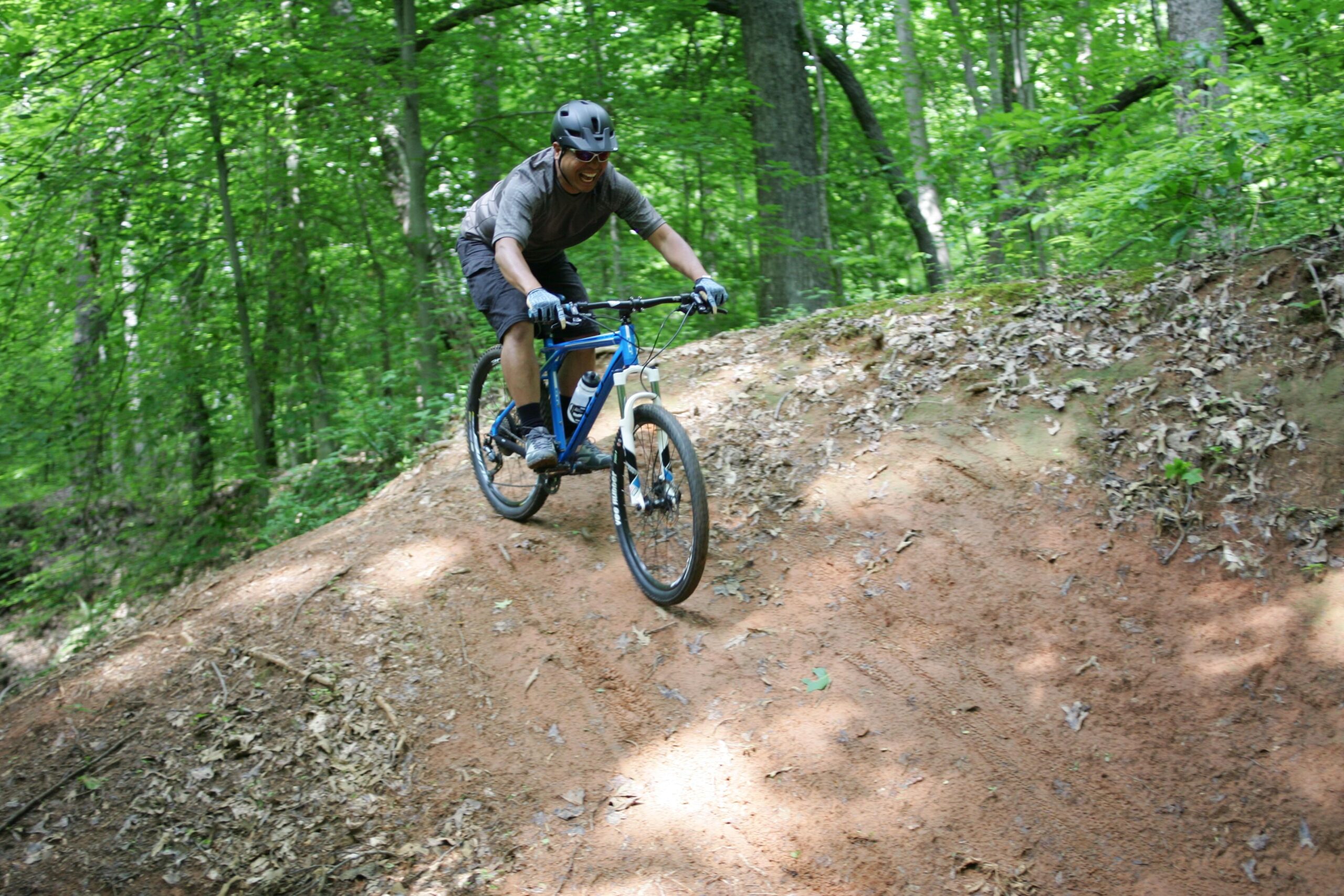 A person riding a mountain bike down a sandy incline surrounded by lush green trees in a forested area. The rider is wearing a helmet and gloves, and appears to be focused on navigating the terrain. Salem Lake mountain bike trail.