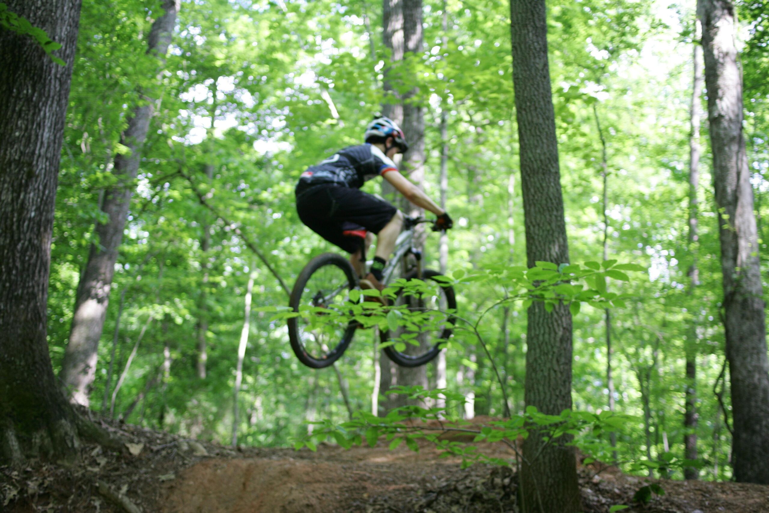 A mountain biker jumping over a dirt ramp in a dense forest, surrounded by green trees and foliage. The rider is wearing a helmet and biking gear, showcasing an active outdoor scene. Salem Lake mountain bike trail.