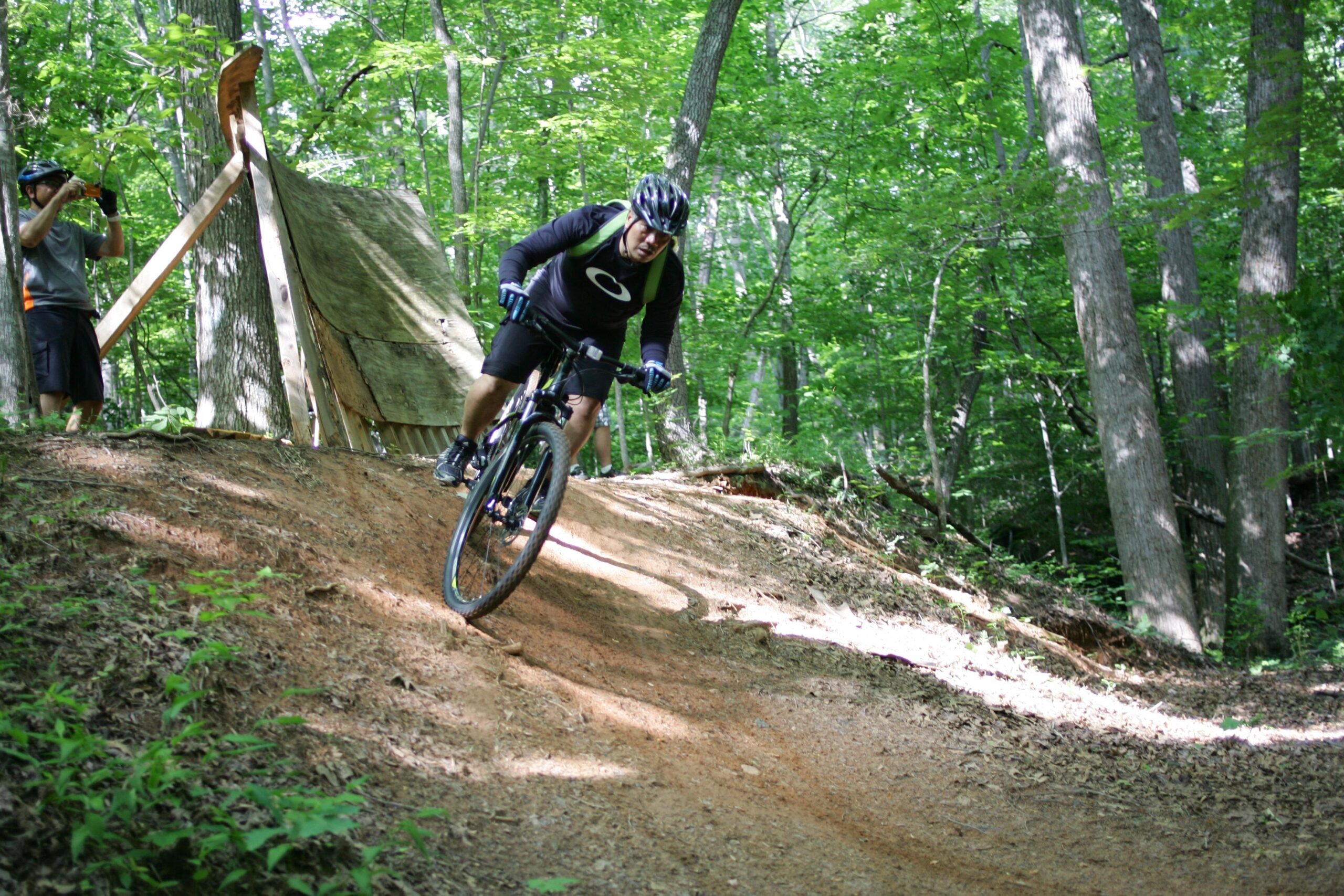 A mountain biker navigating a dirt trail in a forested area, leaning into a turn while riding a bicycle. In the background, another person is taking photos with a camera near a wooden ramp. The scene is surrounded by lush green trees and sunlight filtering through the leaves. Salem Lake mountain bike trail.