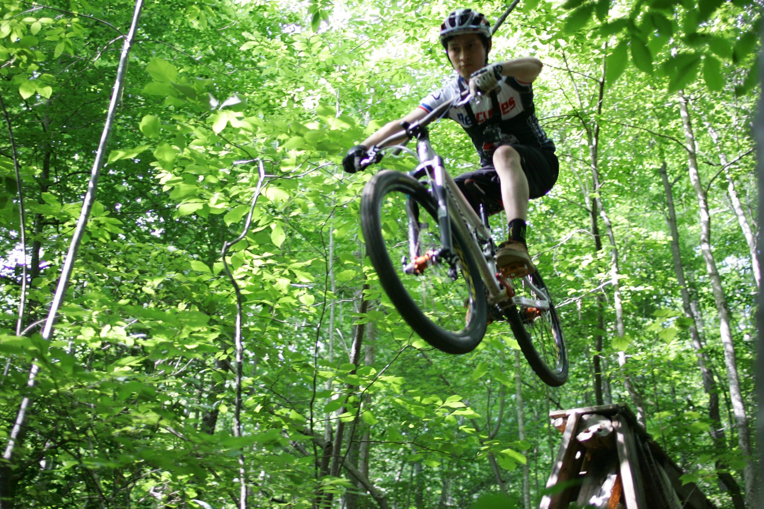 A person riding a mountain bike performing a jump in a lush green forest, surrounded by trees and vibrant foliage. The cyclist is airborne, with a focused expression, wearing a helmet and cycling gear. Salem Lake mountain bike trail.