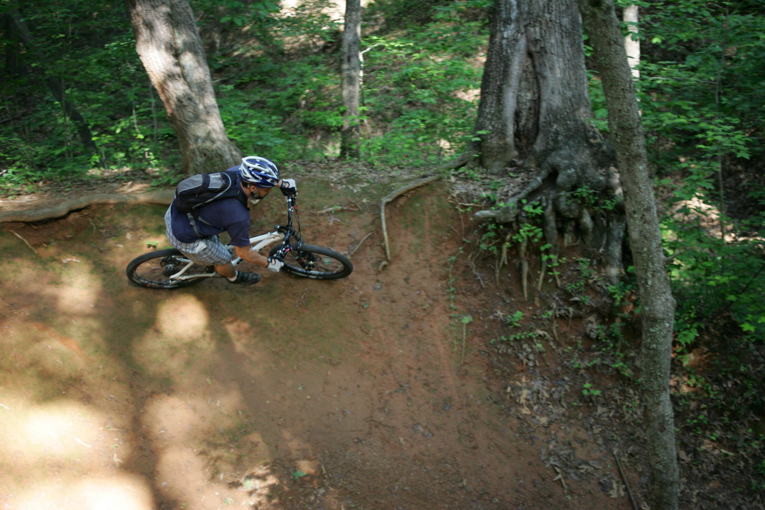 A mountain biker navigating a dirt trail in a forested area, leaning into a turn alongside tree roots and greenery. Salem Lake mountain bike trail.