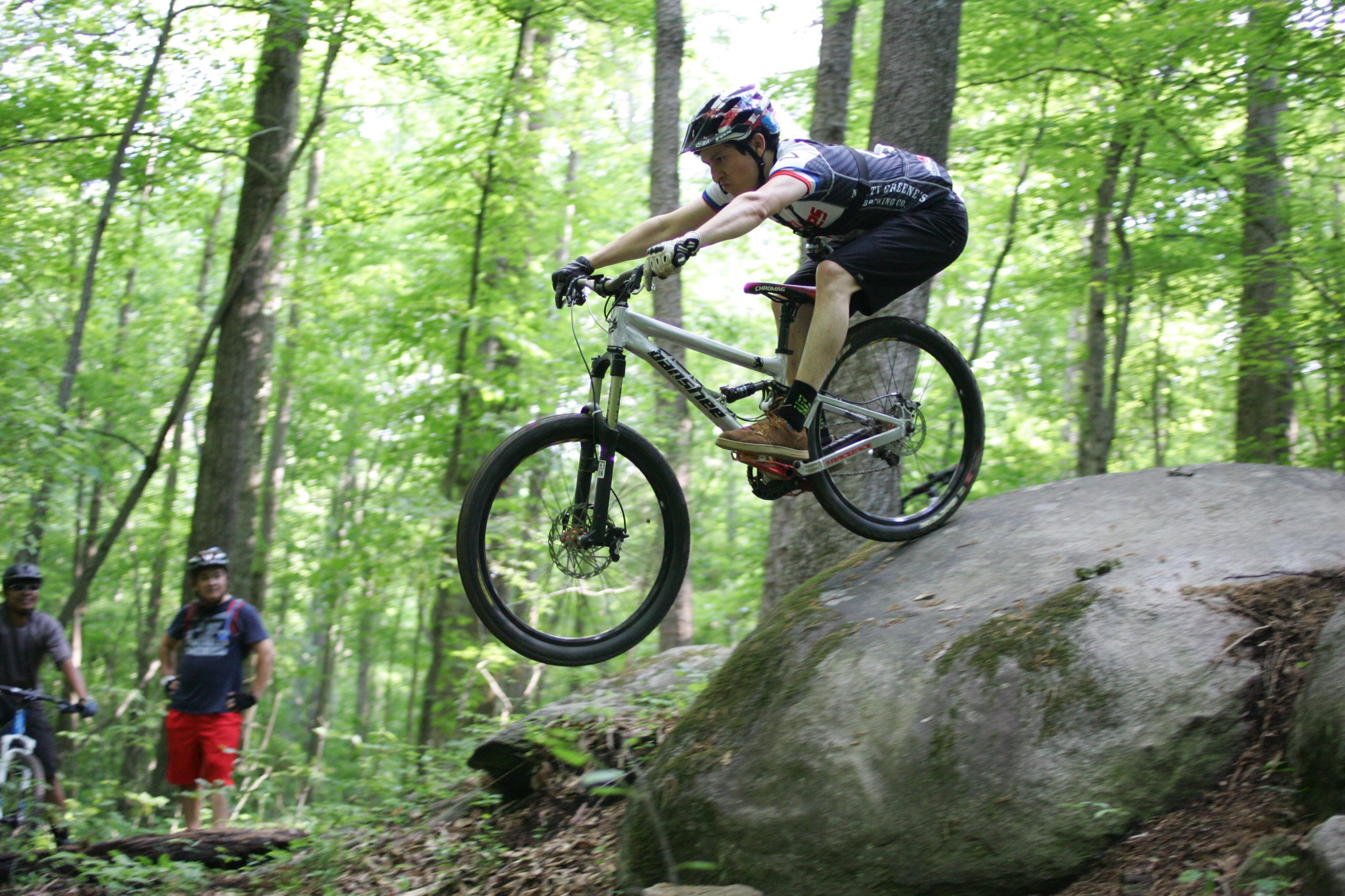 A mountain biker in mid-air, jumping off a large rock on a forested trail, with two observers watching in the background. The scene is surrounded by lush green trees, creating a vibrant outdoor atmosphere. Salem Lake mountain bike trail.