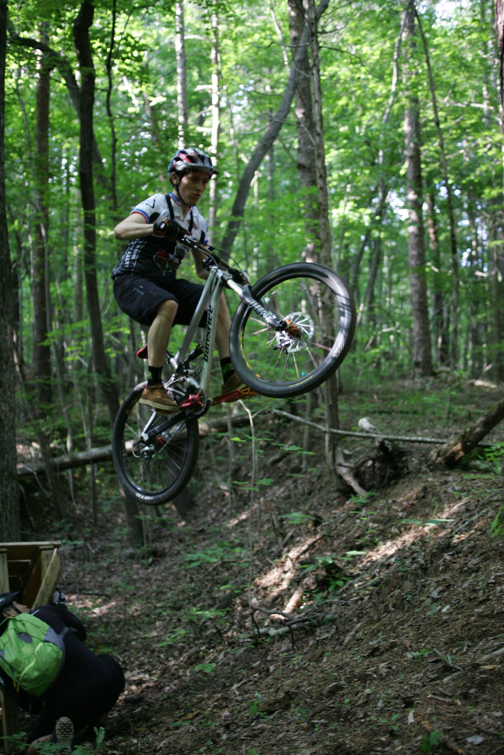 A mountain biker performing a jump in a wooded area, surrounded by green trees and undergrowth. The rider is airborne with one wheel raised off the ground, showcasing an action-packed moment in outdoor cycling. A person in the background is crouching near wooden obstacles on the forest floor. Salem Lake mountain bike trail.