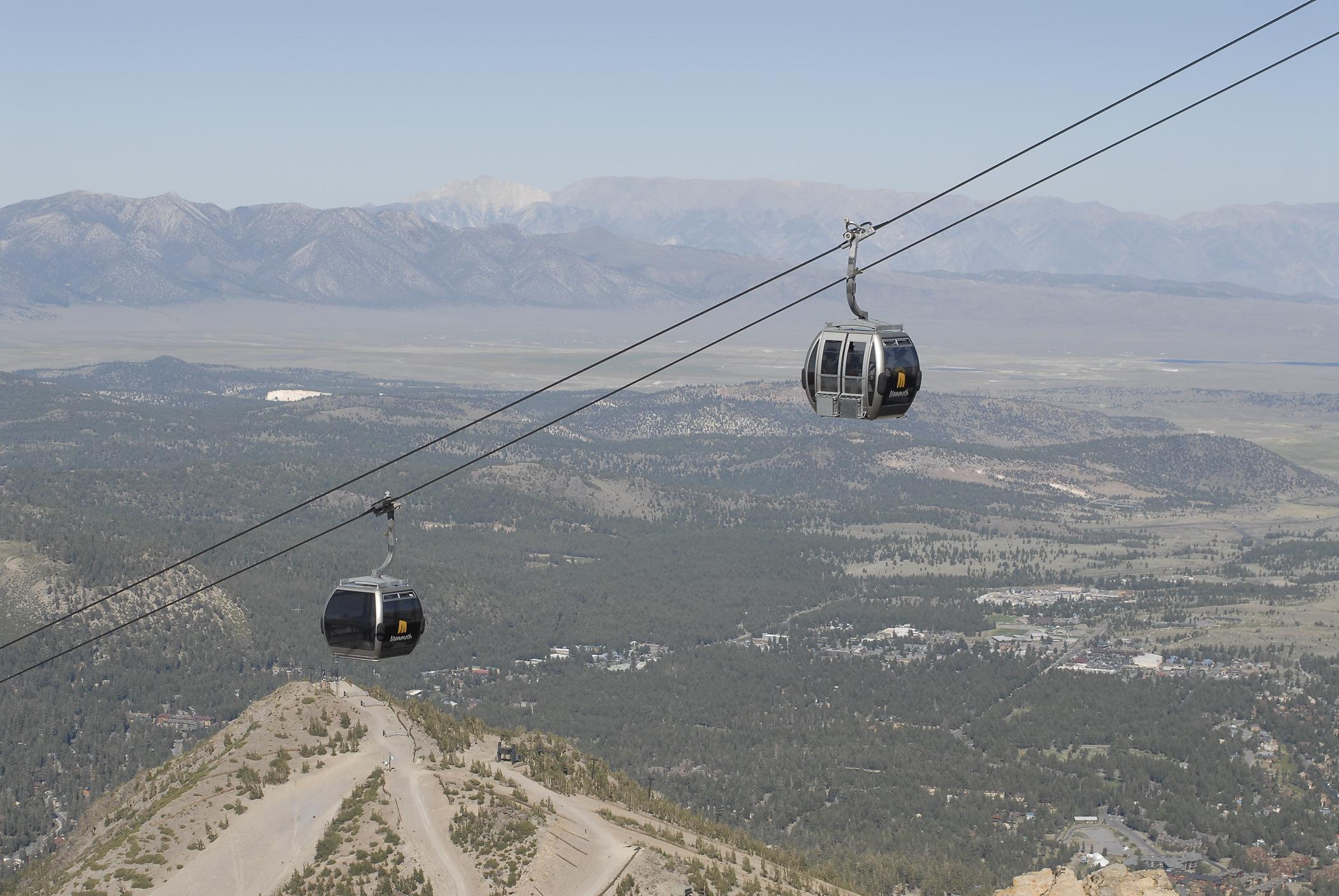 Two gondolas travel along a cable above a mountainous landscape, with lush green forests and a sprawling valley visible below. The background features distant mountains under a clear blue sky. Mammoth Mountain Resort mountain bike trail.