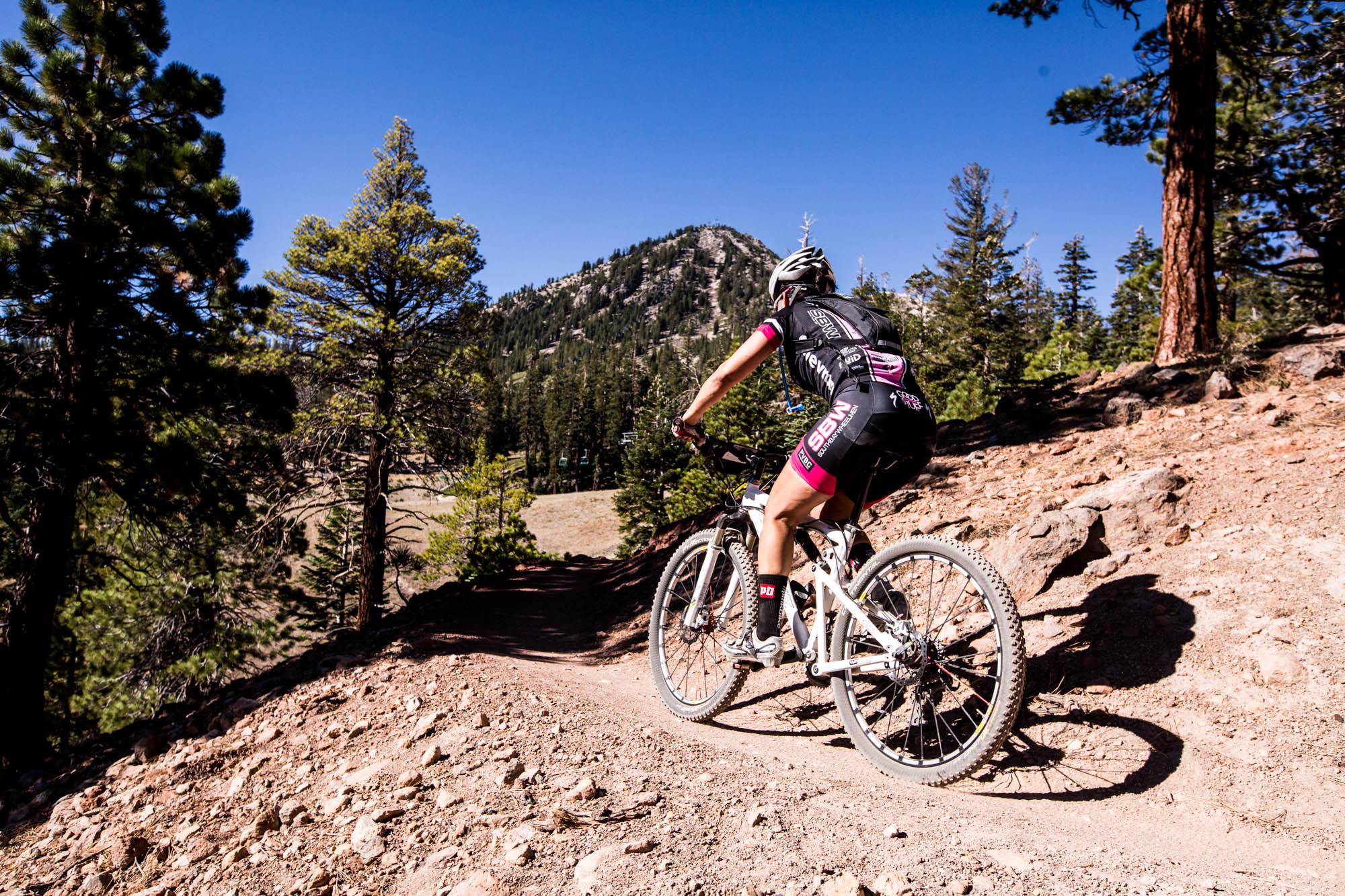 A mountain biker riding along a dirt trail surrounded by pine trees, with a mountain in the background under a clear blue sky. Mammoth Mountain Resort mountain bike trail.