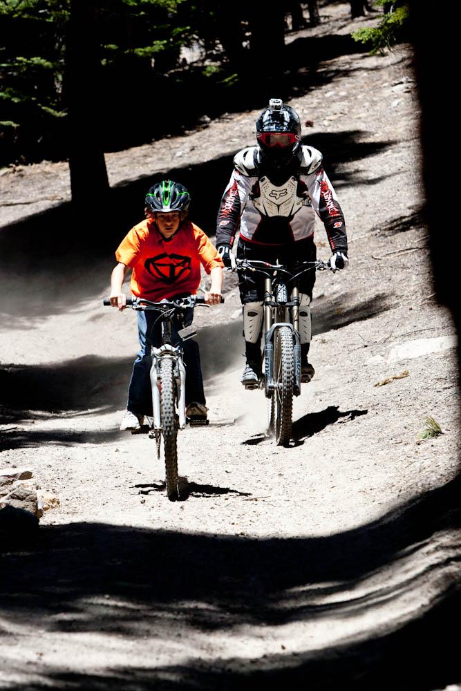 Two cyclists are riding on a dirt trail surrounded by trees. The first rider, a young boy in an orange shirt and green helmet, is pedaling vigorously. The second rider, an adult in a white and black protective outfit and helmet, is keeping pace beside him. Dust is kicking up from the trail, indicating an active biking environment. Mammoth Mountain Resort mountain bike trail.