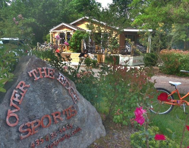 A storefront for "Over the Edge Sports," featuring a rustic building surrounded by greenery and colorful flowers. A large rock sign with the business name is prominently displayed in the foreground, while bicycles and outdoor gear are visible on the porch and around the entrance.