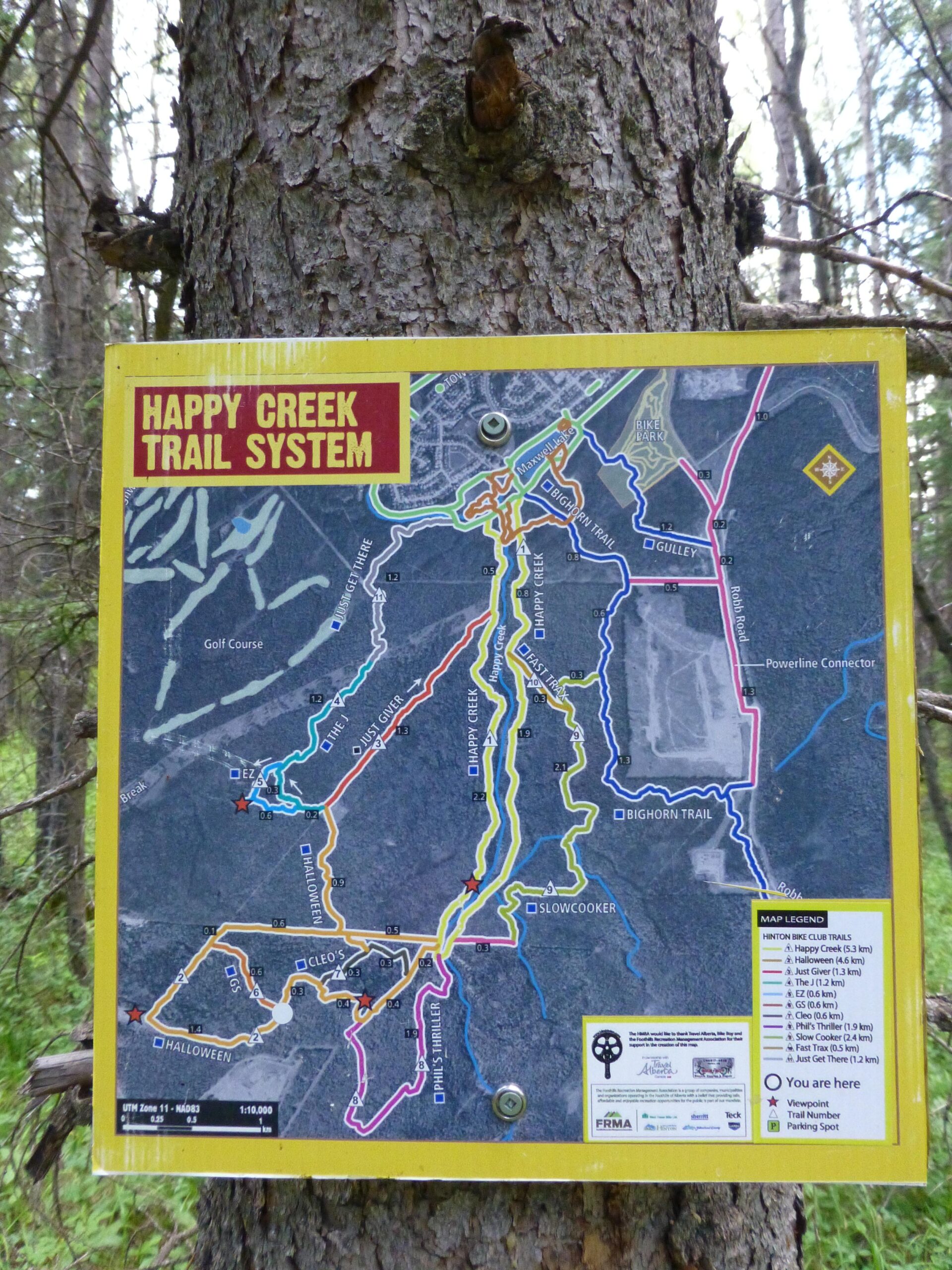 Map of the Happy Creek Trail System, displaying various hiking and biking trails marked in different colors. The map includes trail names, distances, and landmarks, with a location marker indicating "You are here." The sign is mounted on a tree trunk in a wooded area.