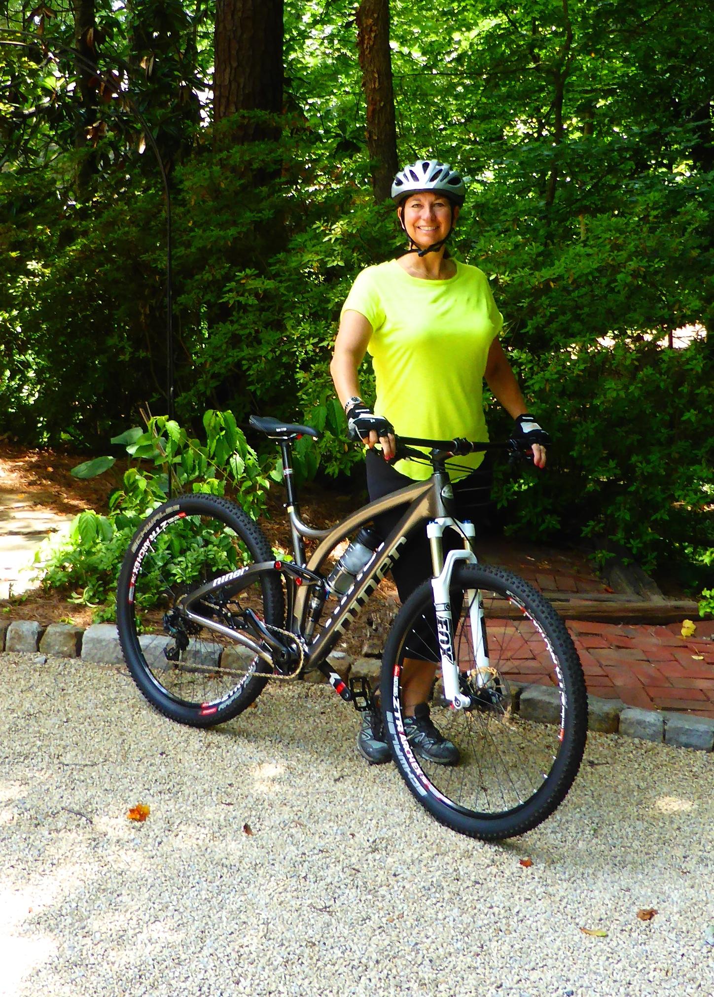 Niner Jet 9 Carbon: A smiling woman wearing a bright yellow shirt and a bicycle helmet stands next to her mountain bike in a lush, green park setting. She is holding onto the bike's handlebars, ready for a ride, with trees and foliage visible in the background.