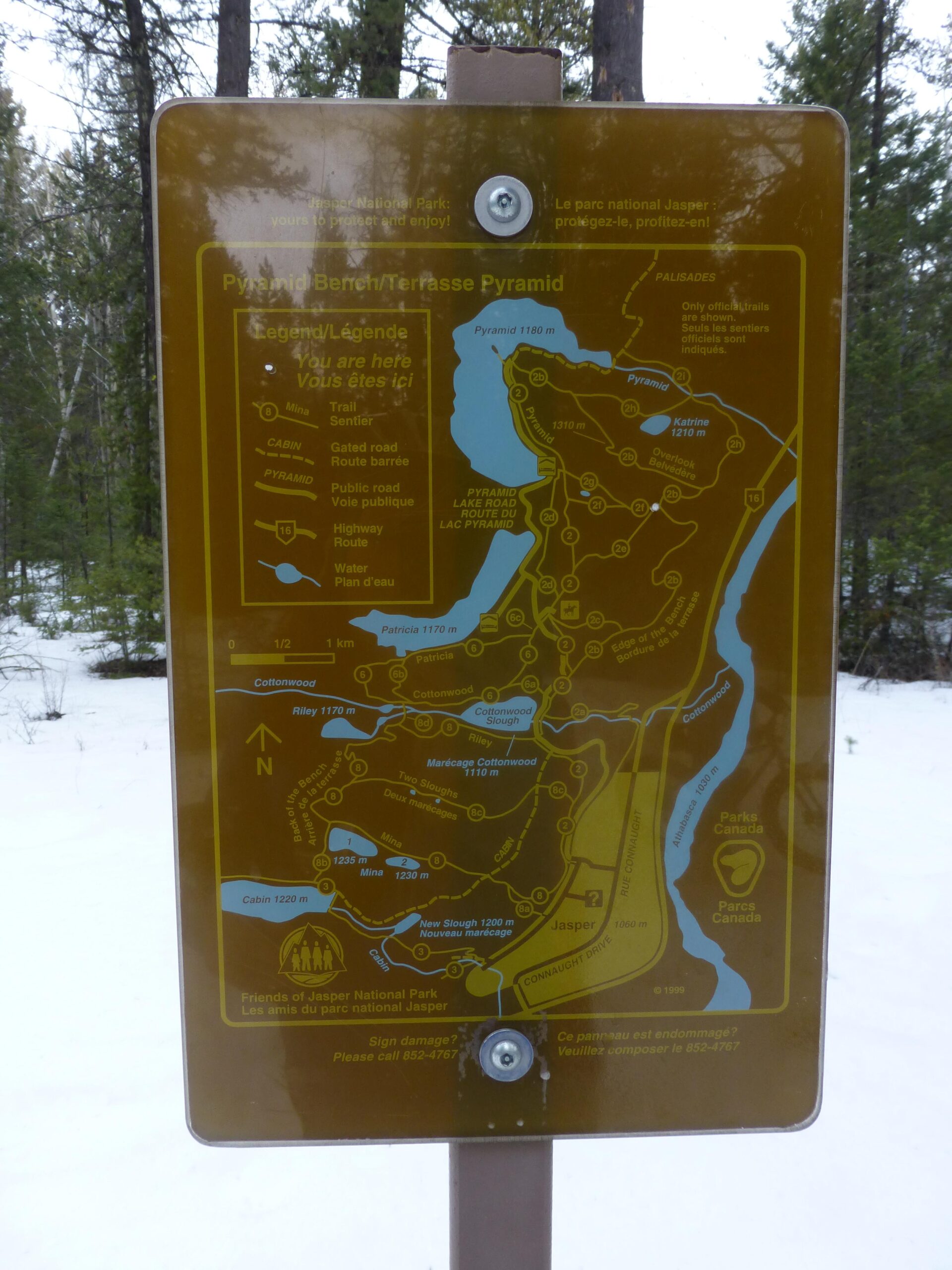 A informational map sign for Pyramid Bench in Jasper National Park, displaying various trails, roads, and landmarks in the area, set against a snowy background. The sign includes details such as elevation markers, trail paths, and a legend in both English and French. Pyramid Bench mountain bike trail.