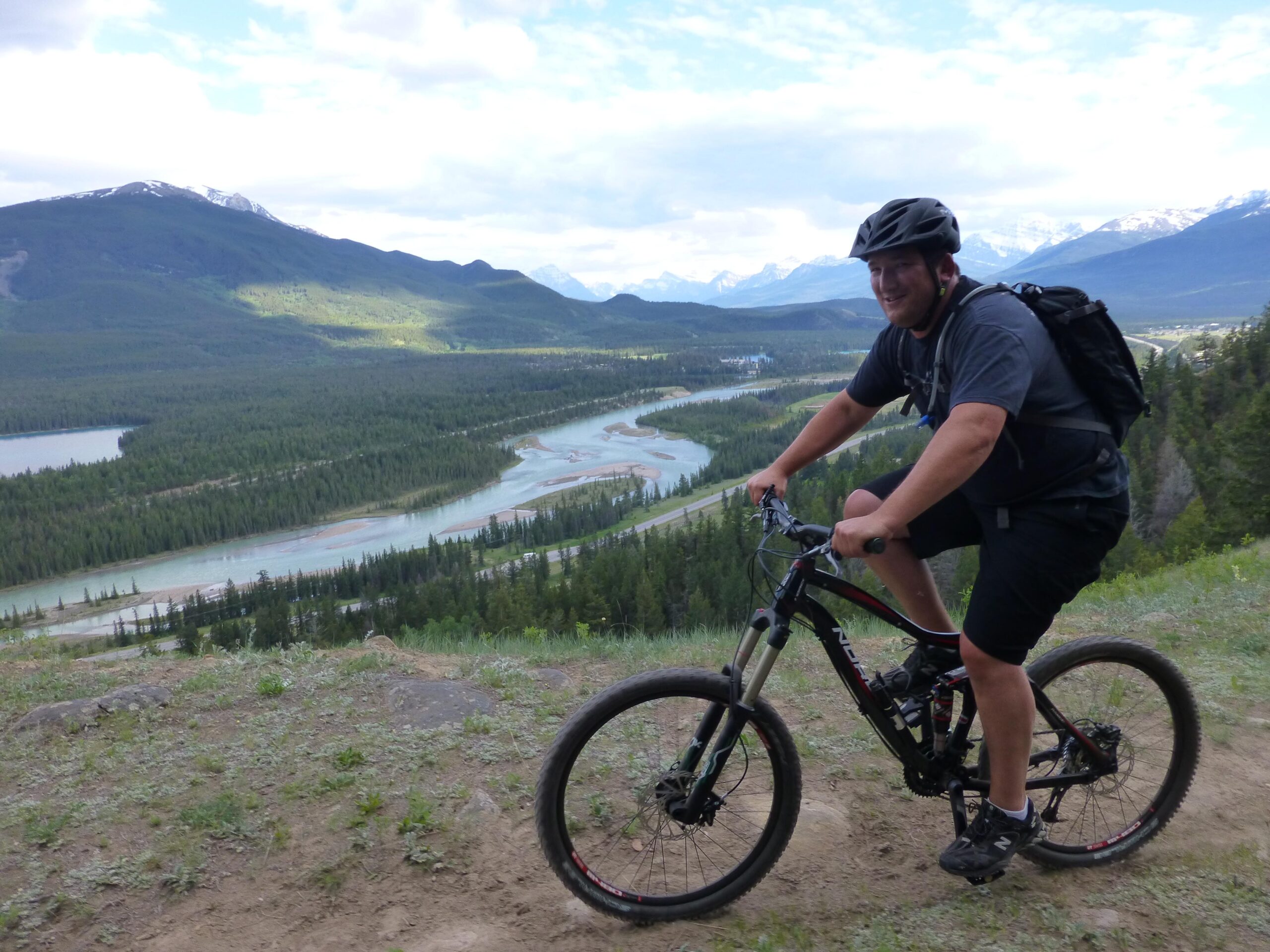 A man in a helmet rides a mountain bike along a dirt path with a scenic view of a river and forested mountains in the background. The landscape features lush greenery and distant snow-capped peaks under a cloudy sky. Pyramid Bench mountain bike trail.