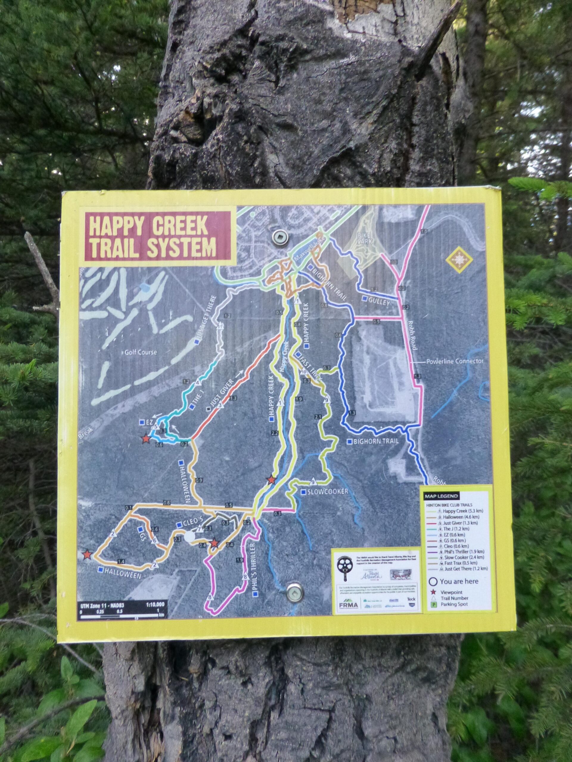 **Alt text:** A detailed trail map of the Happy Creek Trail System, mounted on a tree. The map features various colored trails, landmarks, and a legend indicating trail names and distances. The words "HAPPY CREEK TRAIL SYSTEM" are prominently displayed at the top. Happy Creek Trail System mountain bike trail.