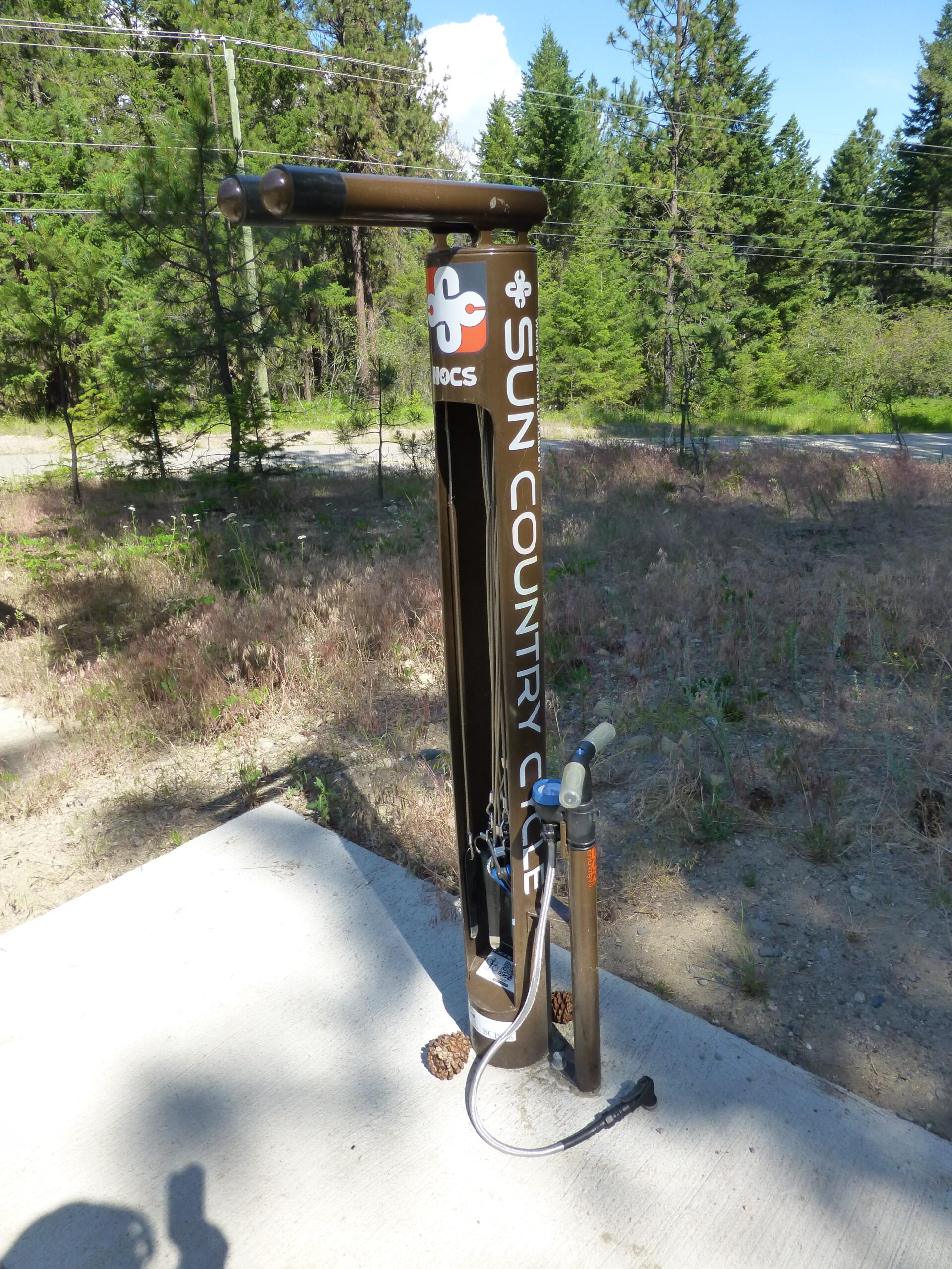 A bicycle repair station featuring a black metal frame with a pump attached, located in a forested area. The station is branded with "SUN COUNTRY CYCLE" and is set on a concrete pad, surrounded by pine trees and underbrush. A hose and tools are available for cyclists' use. Ellison Park mountain bike trail.