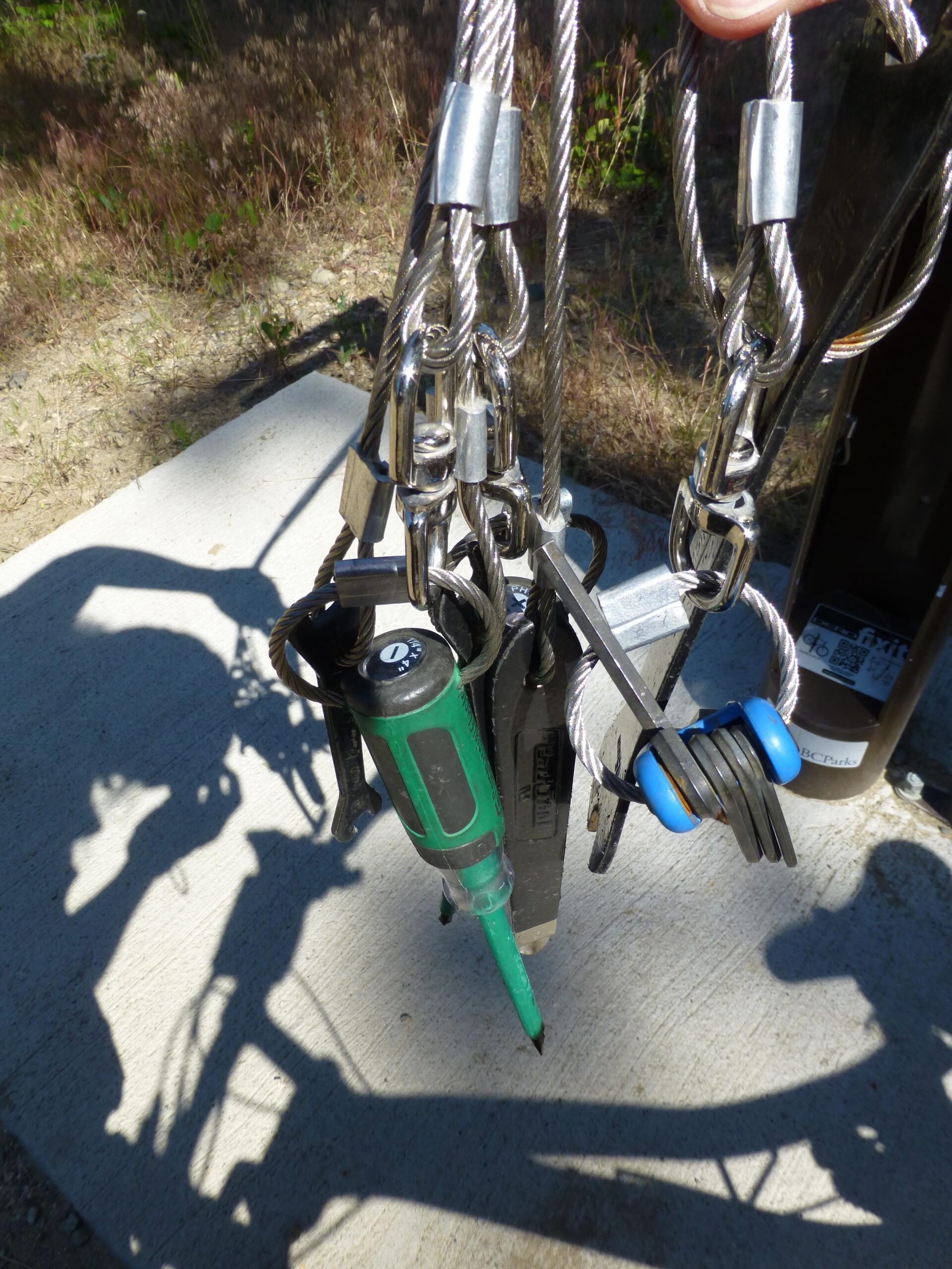 Close-up of a set of keys and tools secured with metal cables, including a green screwdriver and a multi-tool, against a backdrop of a concrete surface and blurred greenery. Ellison Park mountain bike trail.