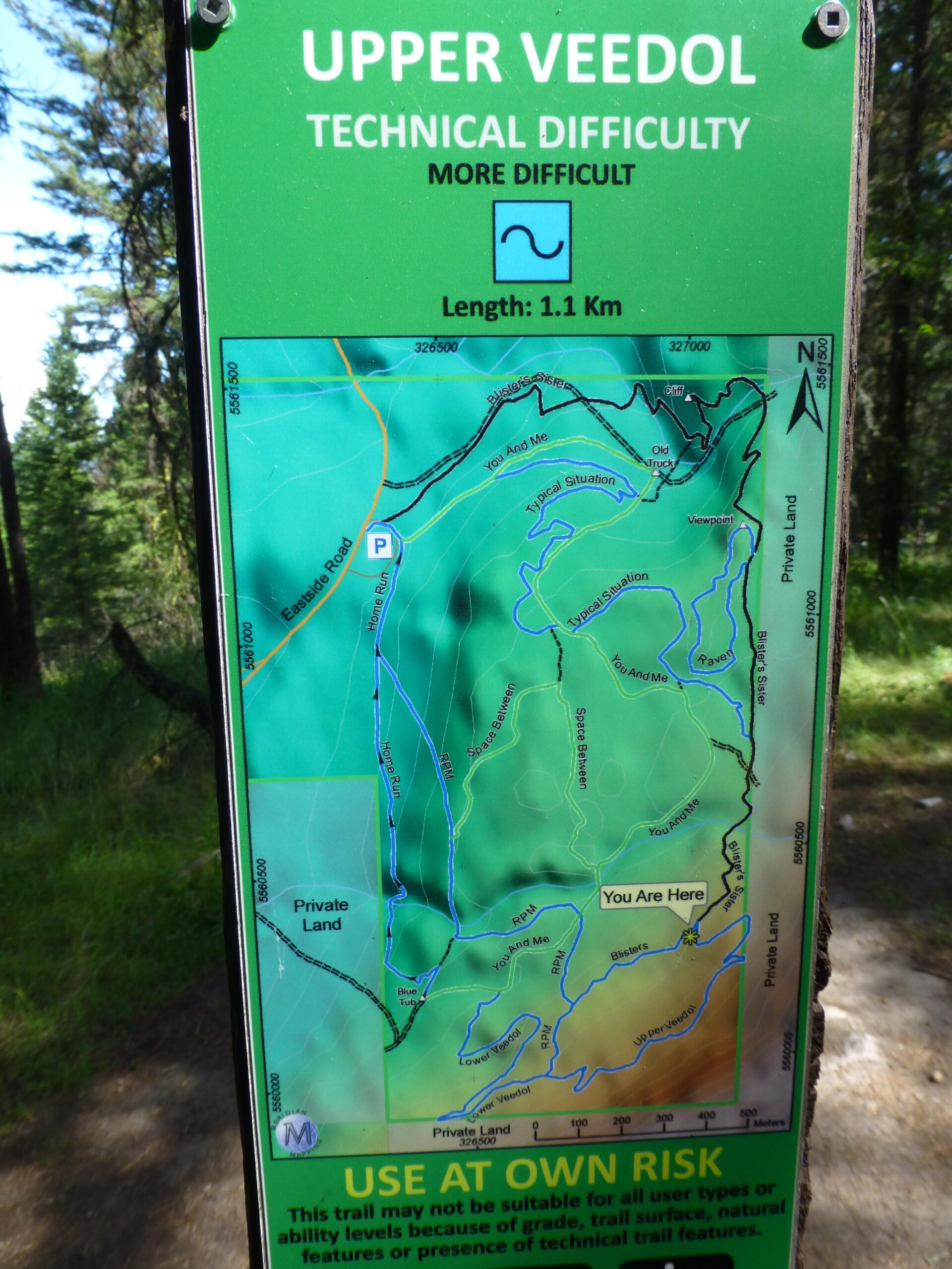 Map of the Upper Veedol trail, indicating a technical difficulty level of "more difficult." The map shows a trail length of 1.1 km, with various route names and a marked "You Are Here" location. Surrounding areas labeled as private land are also indicated, along with a warning to use the trail at one's own risk. The background features trees and natural terrain. Ellison Park mountain bike trail.