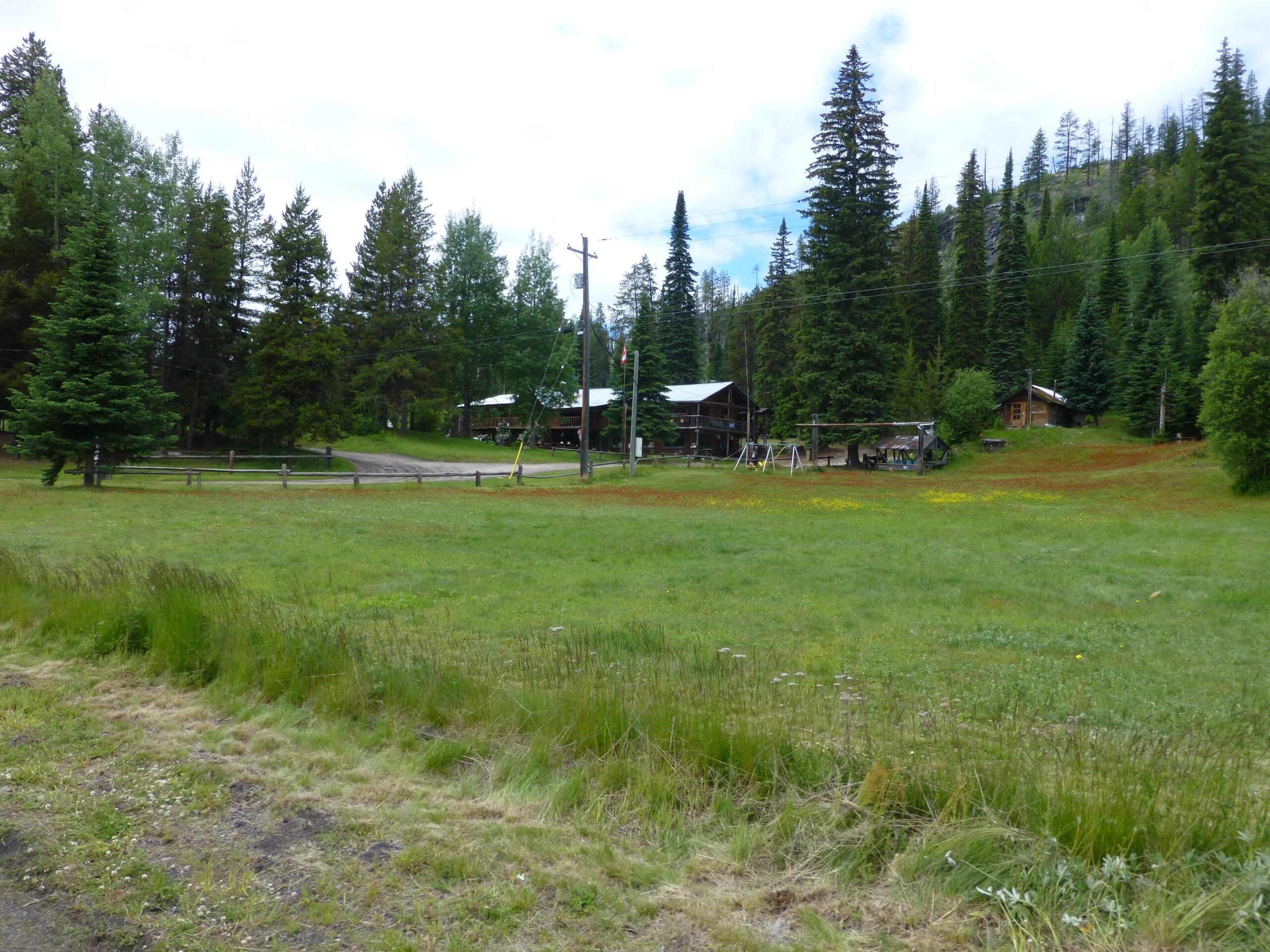 A scenic view of a grassy clearing surrounded by tall trees, with a dirt road leading to a cluster of rustic wooden buildings. In the foreground, there's a field of green grass with patches of wildflowers, and a few swings are visible in the area. The sky is partly cloudy, adding to the serene outdoor atmosphere. Kettle Valley Railway Trail (KVR) Myra Canyon to Penticton return mountain bike trail.