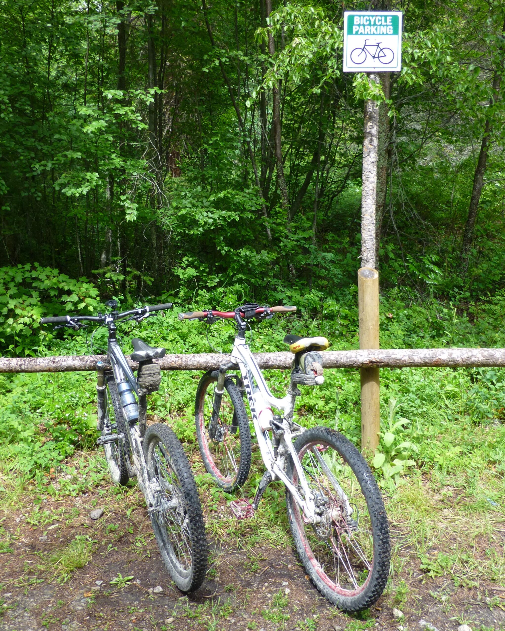 Two mountain bikes are parked against a wooden railing in a lush green forest. A sign reading "BICYCLE PARKING" is displayed nearby, surrounded by trees and foliage. The ground is a mix of dirt and grass, suggesting an outdoor biking trail. Kettle Valley Railway Trail (KVR) Myra Canyon to Penticton return mountain bike trail.