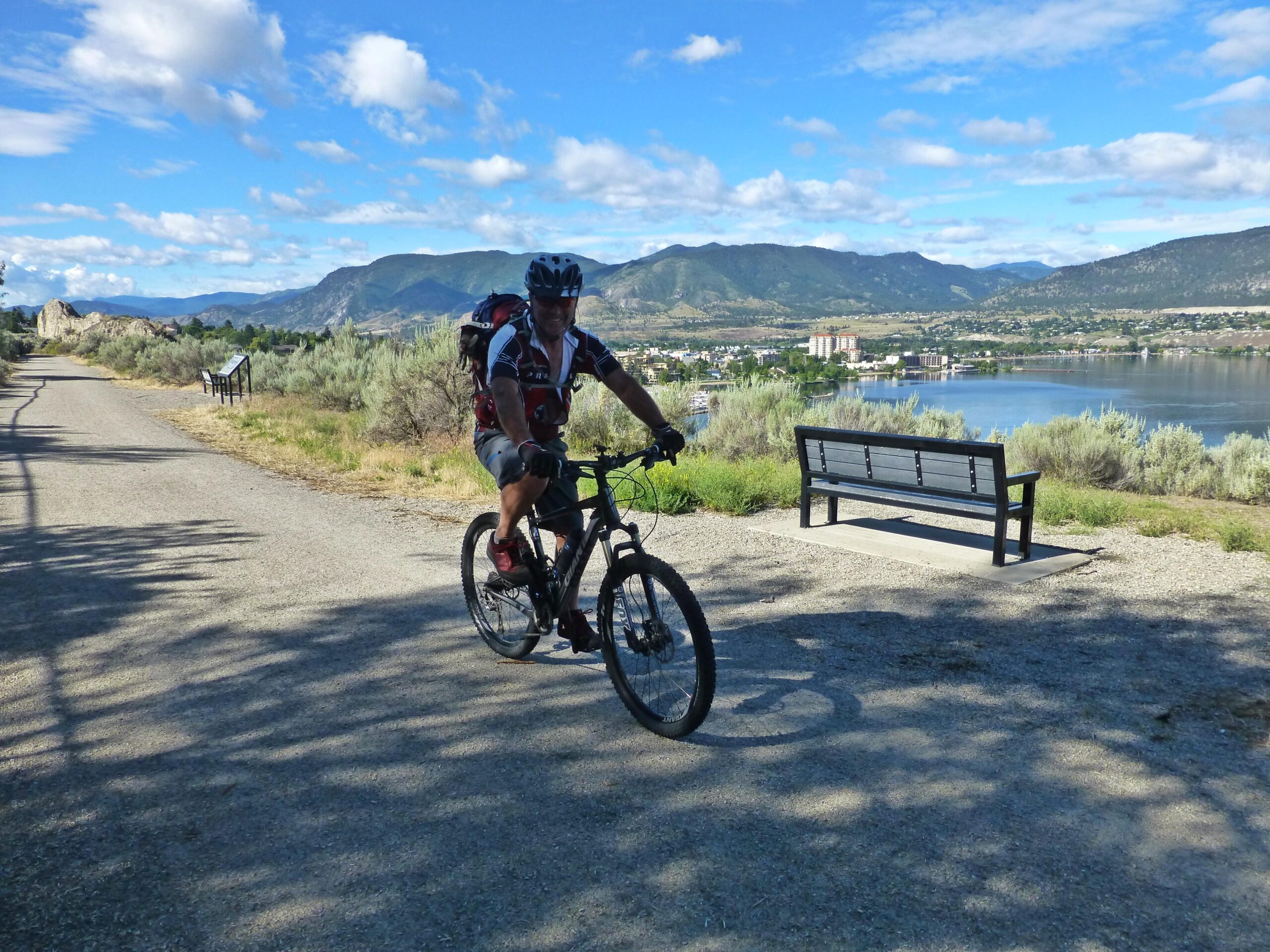 A cyclist rides along a gravel path by a scenic lake, with mountains and blue sky in the background. A wooden bench is positioned nearby, overlooking the water and surrounding landscape. Kettle Valley Railway Trail (KVR) Myra Canyon to Penticton return mountain bike trail.