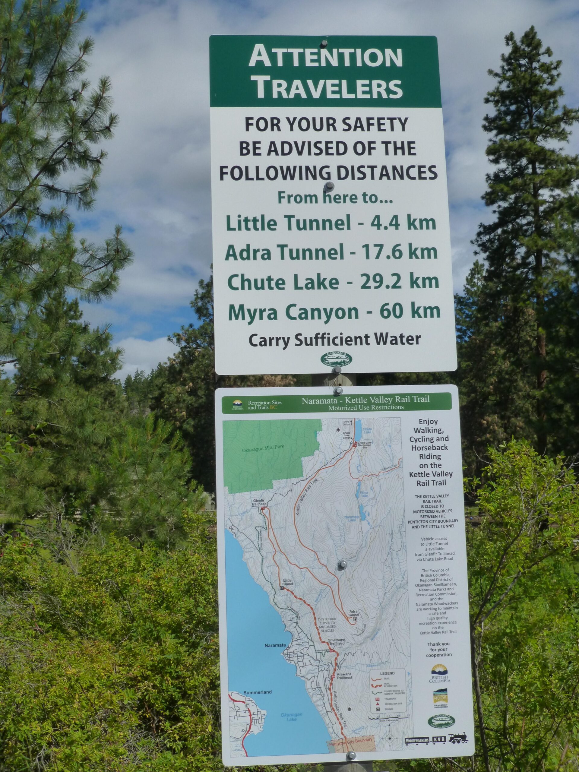 A green and white information sign for travelers displaying distances to nearby attractions. The sign lists distances in kilometers to Little Tunnel (4.4 km), Adra Tunnel (17.6 km), Chute Lake (29.2 km), and Myra Canyon (60 km). It includes a reminder to carry sufficient water and features a map of the Naramata-Kettle Valley Rail Trail area, highlighting routes for walking, cycling, and horseback riding. Pine trees and a cloudy sky are visible in the background. Kettle Valley Railway Trail (KVR) Myra Canyon to Penticton return mountain bike trail.