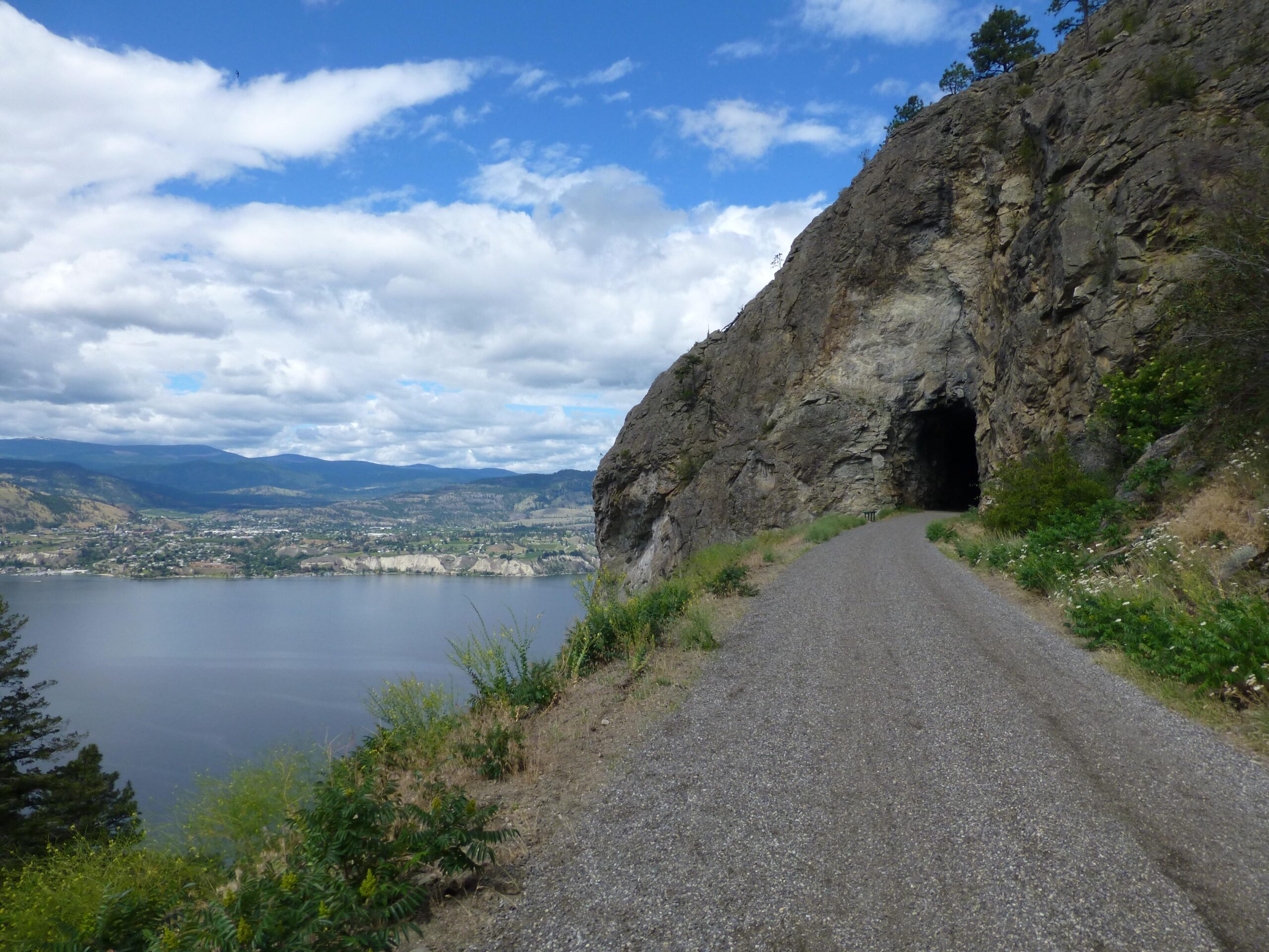 A gravel path leads towards a dark tunnel carved into a rocky cliff, with a serene lake visible on the left and rolling hills in the background under a partly cloudy sky. Kettle Valley Railway Trail (KVR) Myra Canyon to Penticton return mountain bike trail.