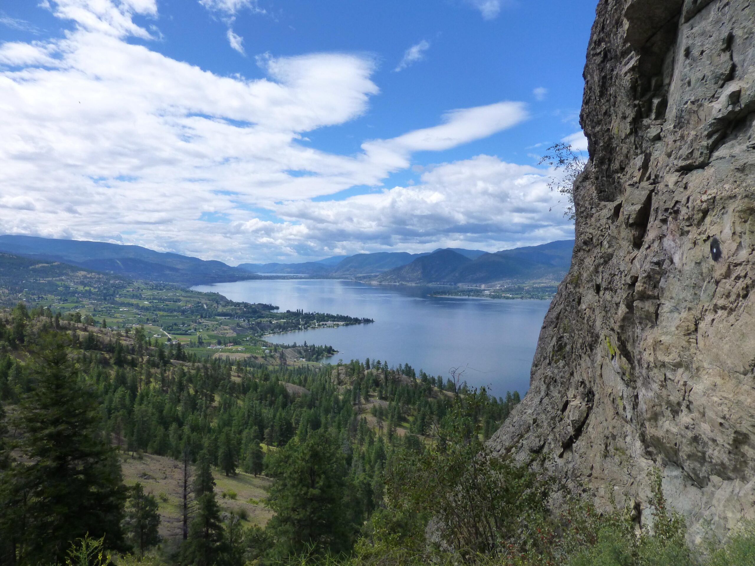 A scenic view of a lake surrounded by lush greenery and rolling hills, with mountains in the background under a partly cloudy sky. The foreground features a rocky cliff, adding depth to the landscape. Kettle Valley Railway Trail (KVR) Myra Canyon to Penticton return mountain bike trail.