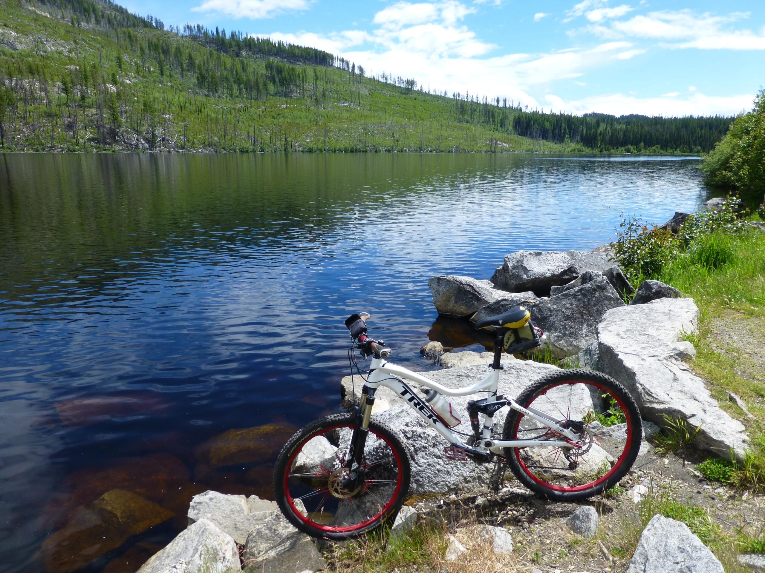 A mountain bike parked on a rocky shore beside a calm lake, surrounded by lush green hills and scattered trees under a blue sky with fluffy clouds. The water reflects the landscape, creating a serene outdoor scene. Kettle Valley Railway Trail (KVR) Myra Canyon to Penticton return mountain bike trail.