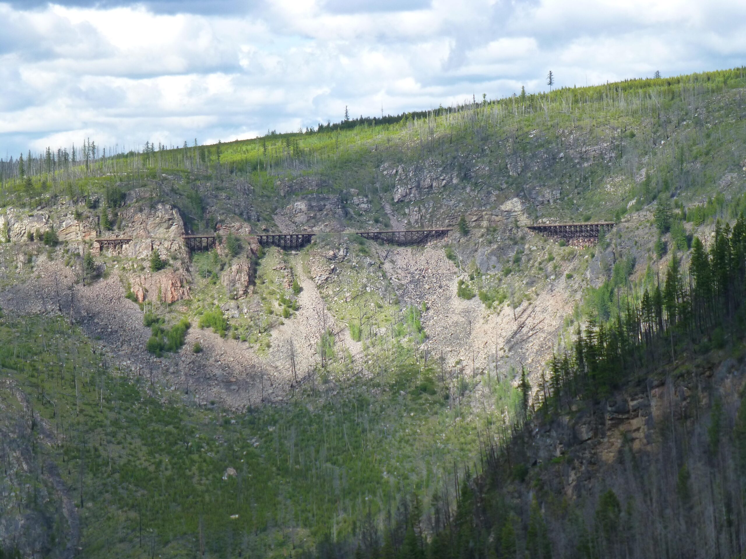 A scenic view of a mountainous landscape featuring rocky hills and patches of greenery. In the background, an old railway trestle bridge runs along the slope, set against a cloudy sky. The foreground shows uneven terrain with scattered vegetation and remnants of trees, highlighting the natural beauty of the area. Kettle Valley Railway Trail (KVR) Myra Canyon to Penticton return mountain bike trail.