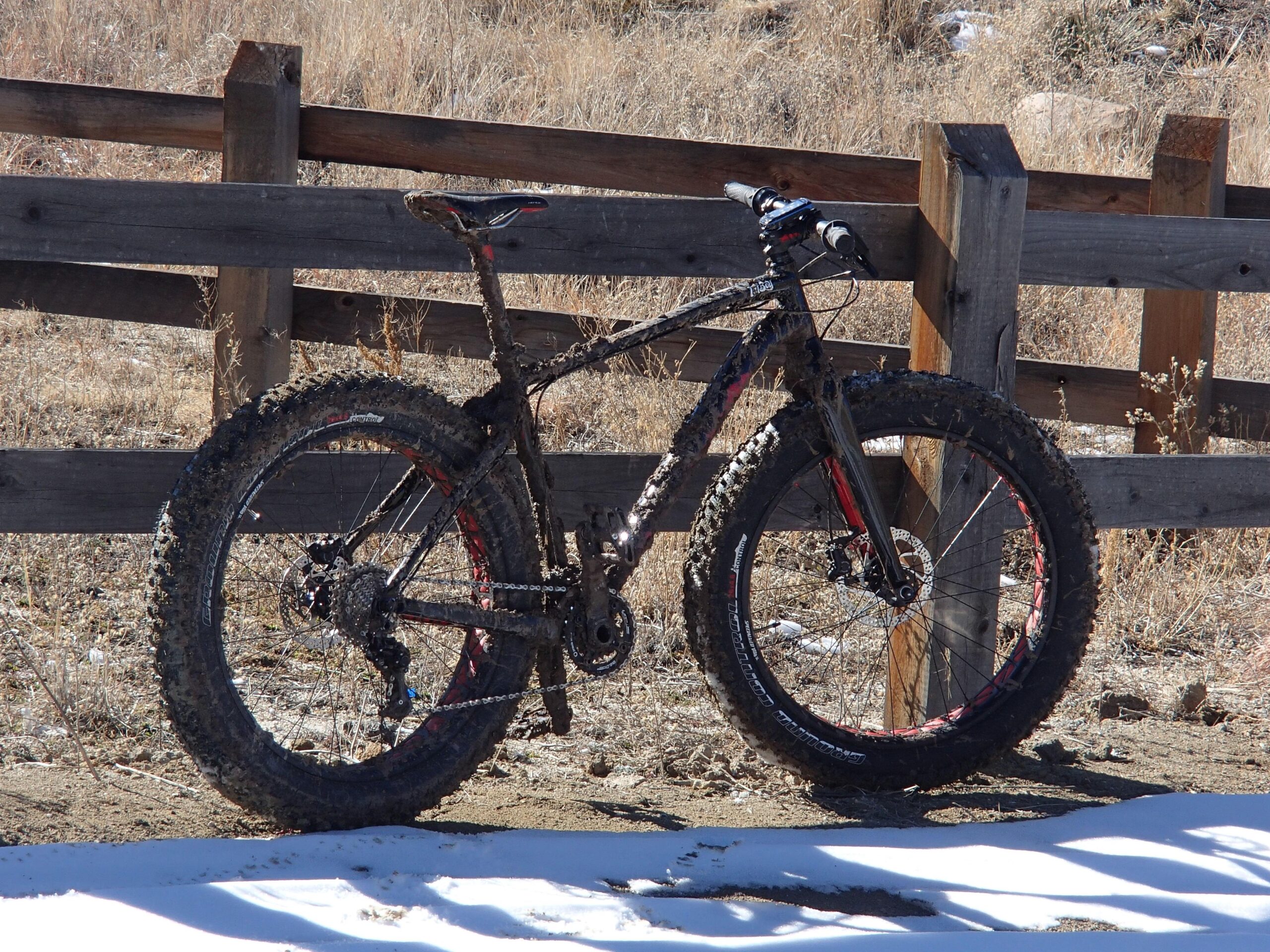 Specialized Fatboy Expert: A muddy fat tire mountain bike leaning against a wooden fence, with dry grass and patches of snow in the background.