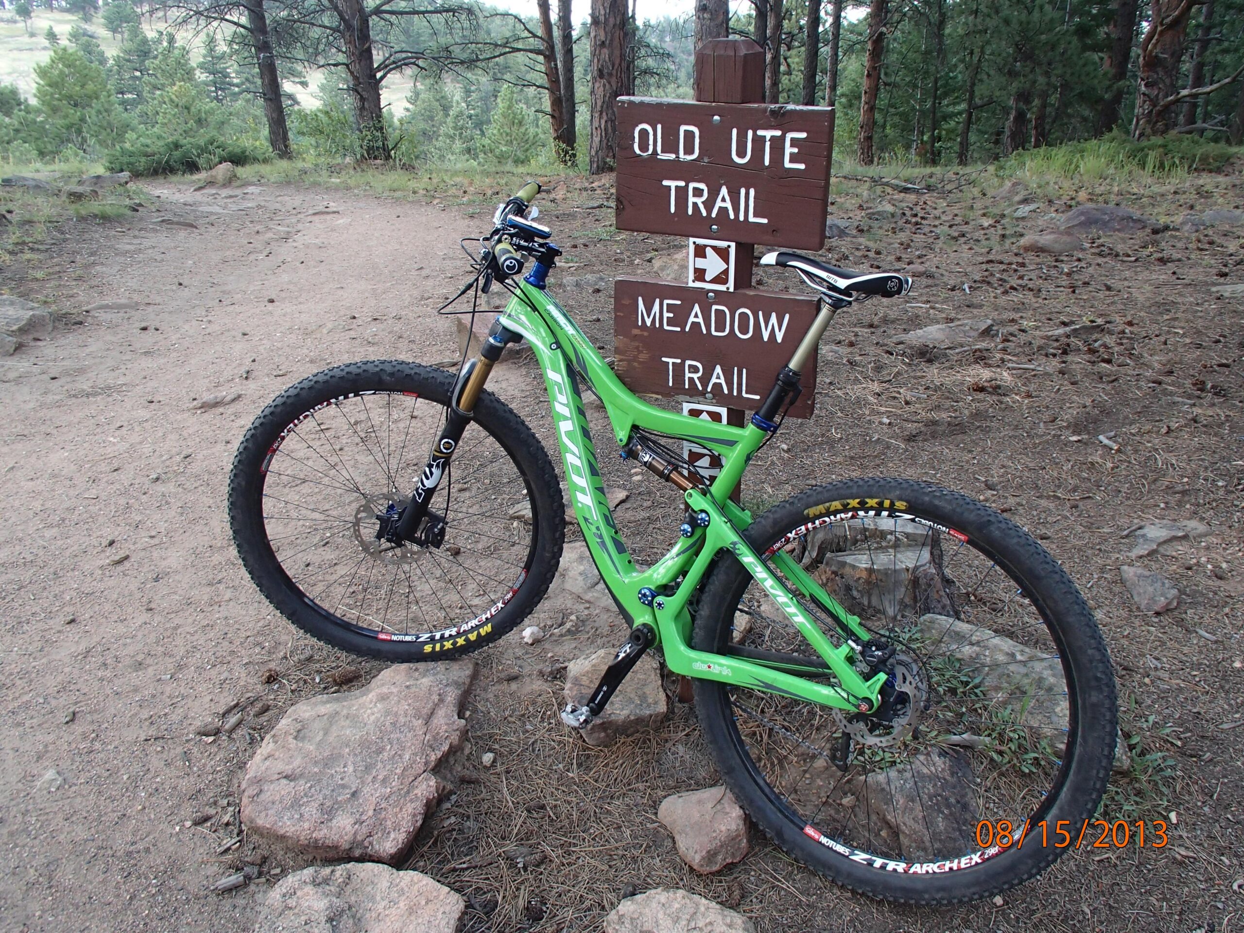Pivot Mach 429: A green mountain bike parked next to a trail sign indicating the directions for "Old Ute Trail" and "Meadow Trail," surrounded by pine trees and a gravel path.