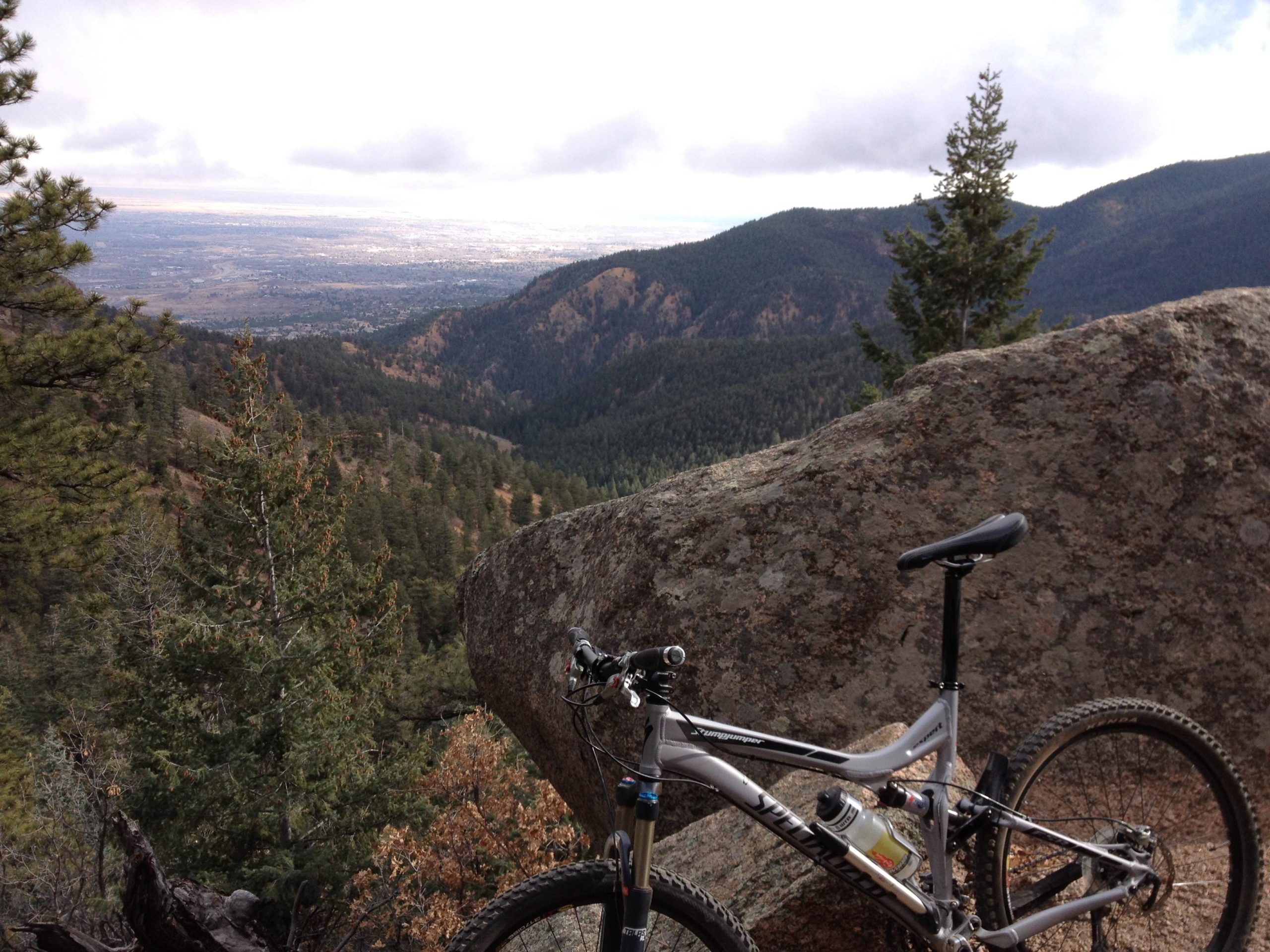 Mountain bike leaning against a large rock with a scenic view of a valley and distant mountains under a partly cloudy sky. Pine trees dot the landscape, emphasizing the natural surroundings. Palmer Trail / Section 16 mountain bike trail.