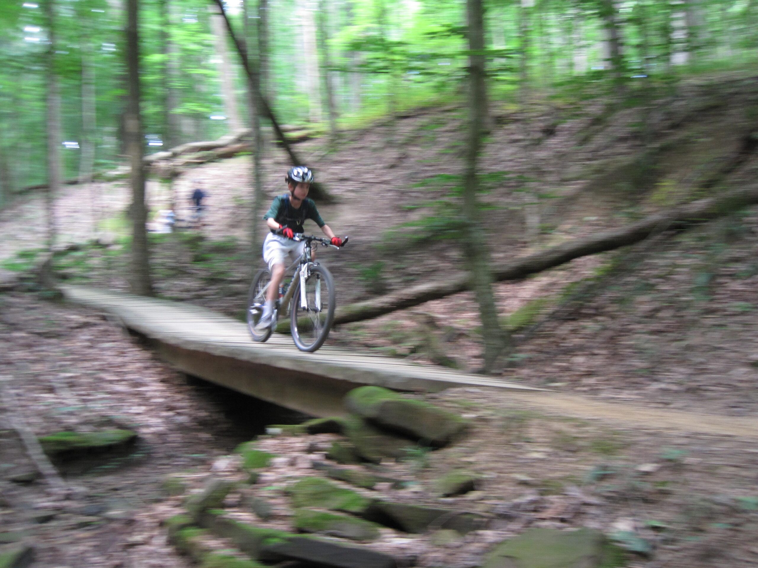 A child riding a mountain bike over a wooden bridge in a wooded area, surrounded by green trees and underbrush, with motion blur conveying speed. Brown County Park mountain bike trail.