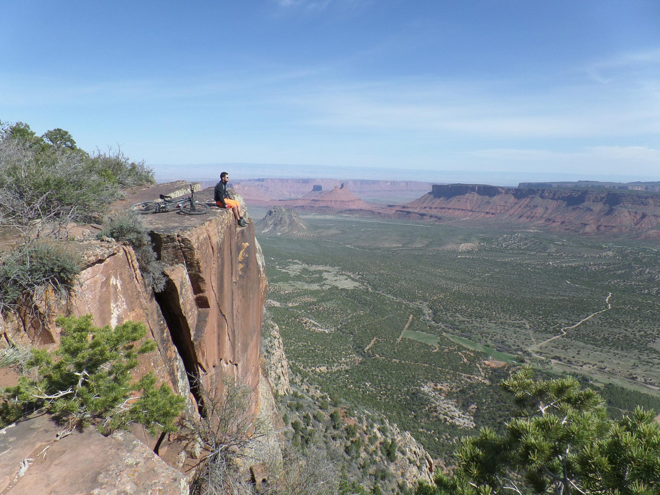 A cyclist sitting on the edge of a rocky cliff, overlooking a vast landscape of red rock formations and green hills under a clear blue sky. A mountain bike is propped up nearby. The Whole Enchilada mountain bike trail.