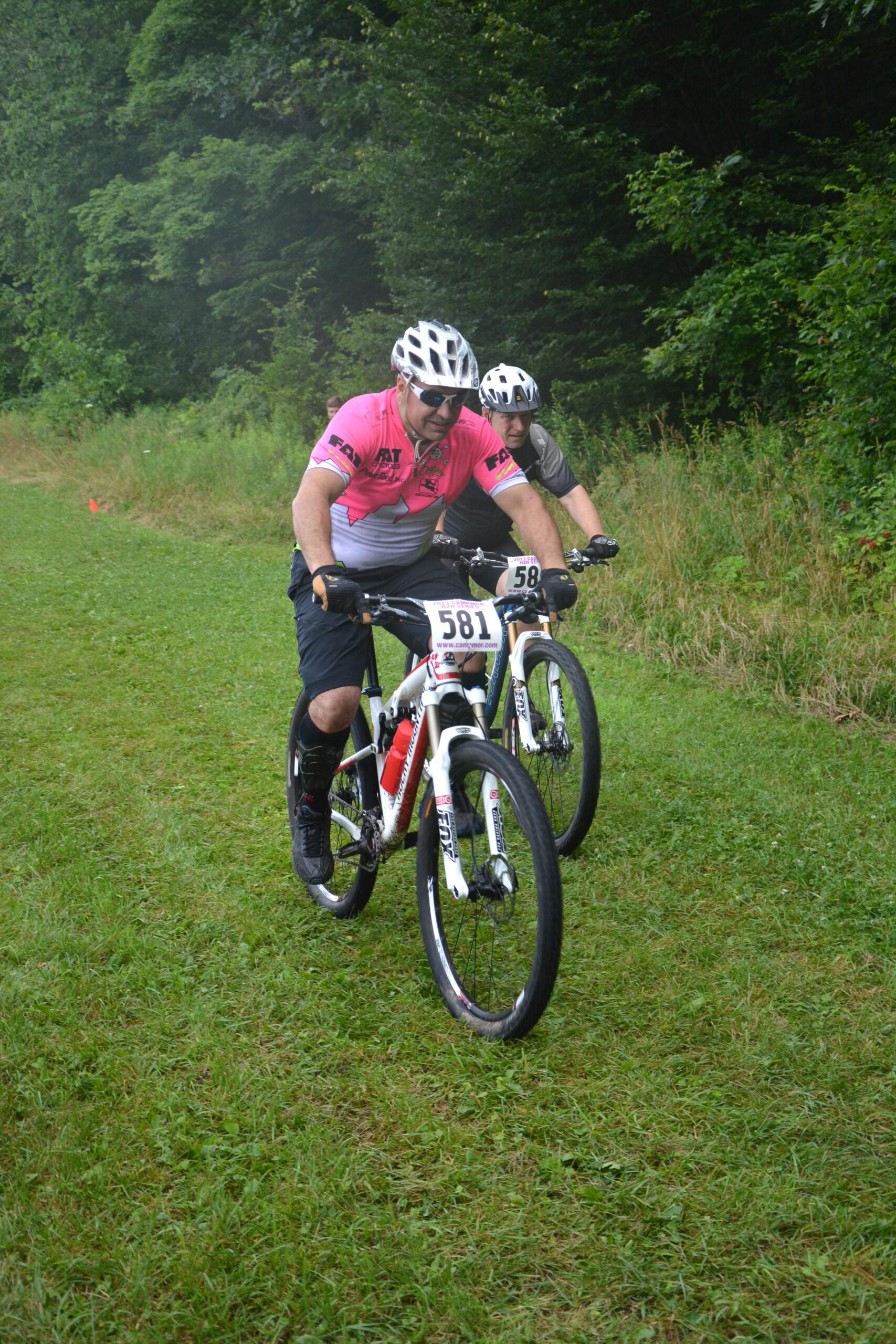 Rocky Mountain Instinct 950: Two mountain bikers pedal through a grassy trail surrounded by trees. One rider is wearing a pink jersey with the number 581, while the other has a gray jersey with the number 58. Both cyclists are focused on the path ahead, showcasing their passion for the sport in a natural setting.