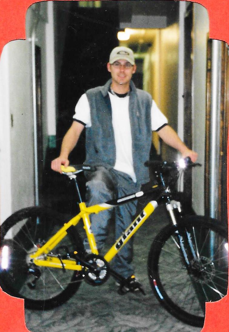 Giant Ranier: A man wearing a gray vest and a baseball cap poses next to a bright yellow mountain bike in a hallway. The background features light-colored walls and a carpeted floor. The image captures a casual moment, highlighting the bike and the man's relaxed expression.