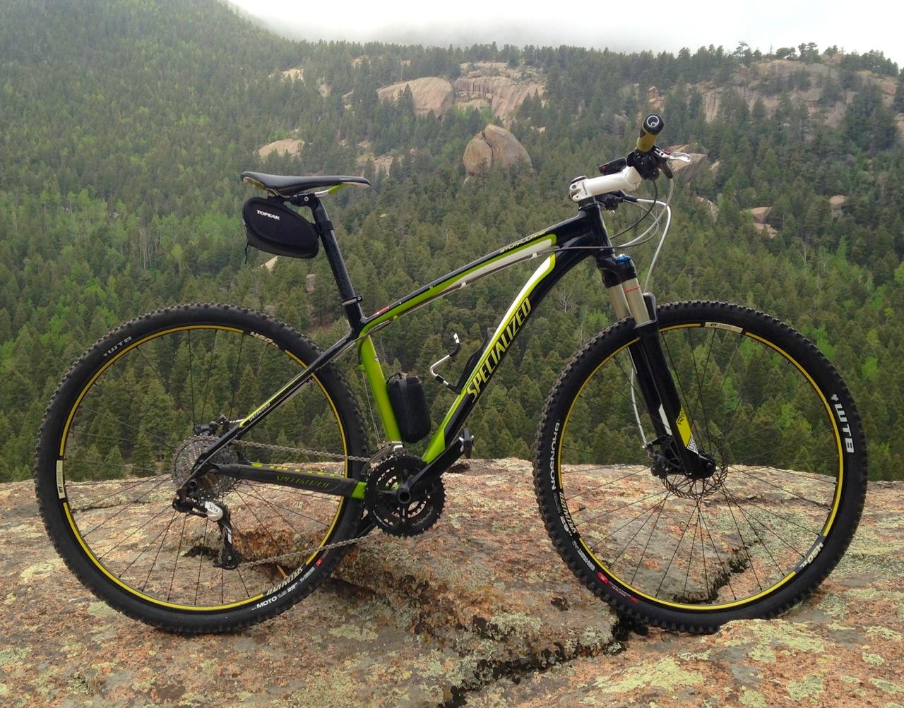 Specialized Stumpjumper Comp 29er: A mountain bike parked on a rocky surface overlooking a lush green forest and distant mountains under a cloudy sky. The bike features a black and green frame with yellow accents, suitable for off-road trails.