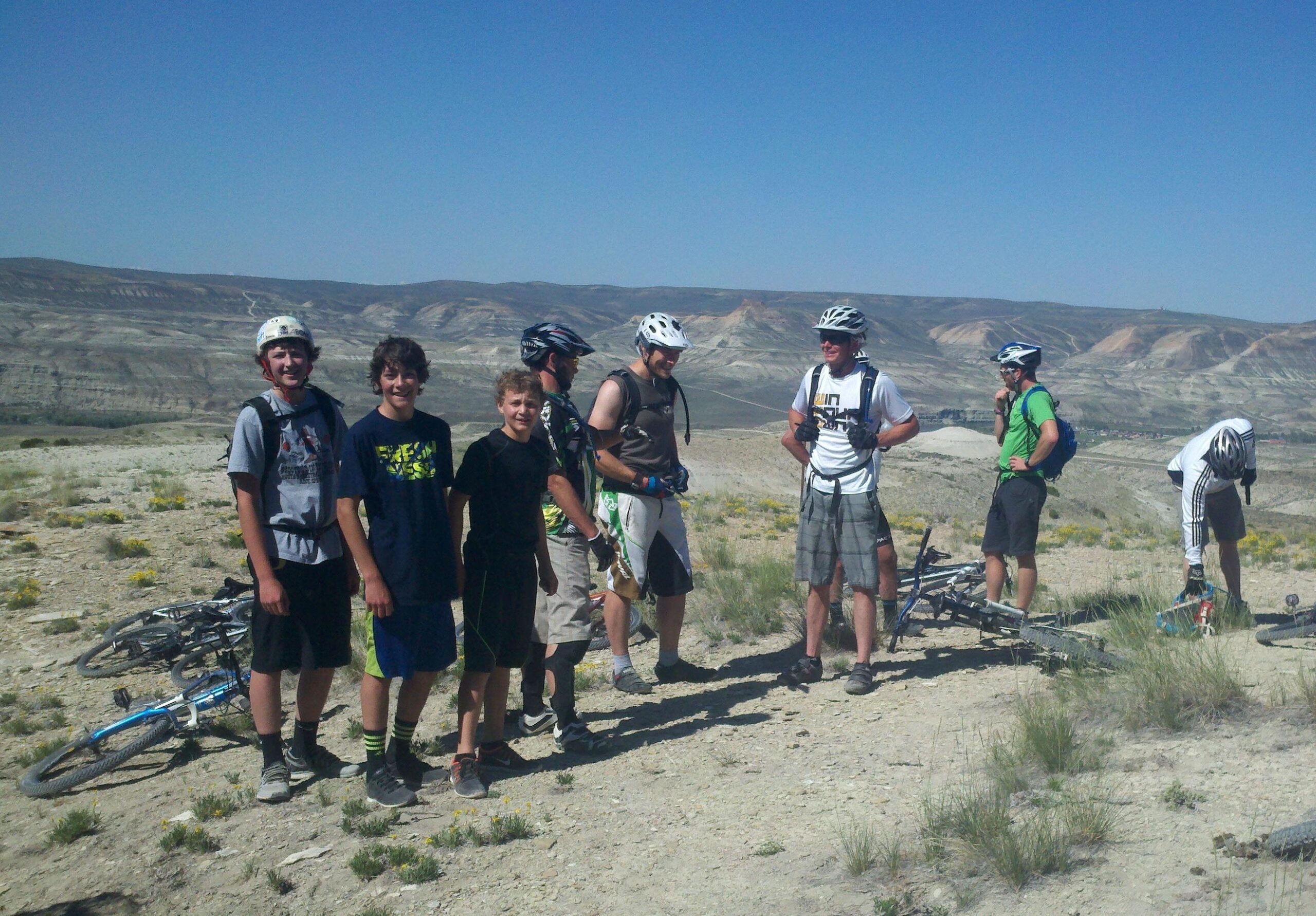 A group of six individuals, including young teenagers and adults, are gathered on a scenic hillside during a mountain biking trip. They are wearing helmets and biking gear, with bicycles resting nearby. The background features a rugged landscape with rolling hills and blue skies. The group appears to be taking a break, smiling and interacting with one another. Wilkins Peak Trails mountain bike trail.