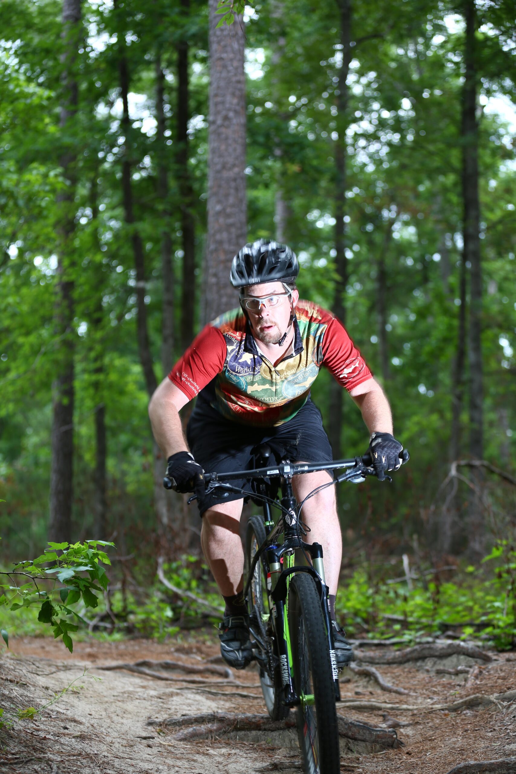 A mountain biker wearing a colorful jersey and helmet rides on a dirt path through a wooded area, surrounded by green trees. The cyclist is focused and navigating over roots and uneven terrain. Forks Area Trail System (FATS) mountain bike trail.